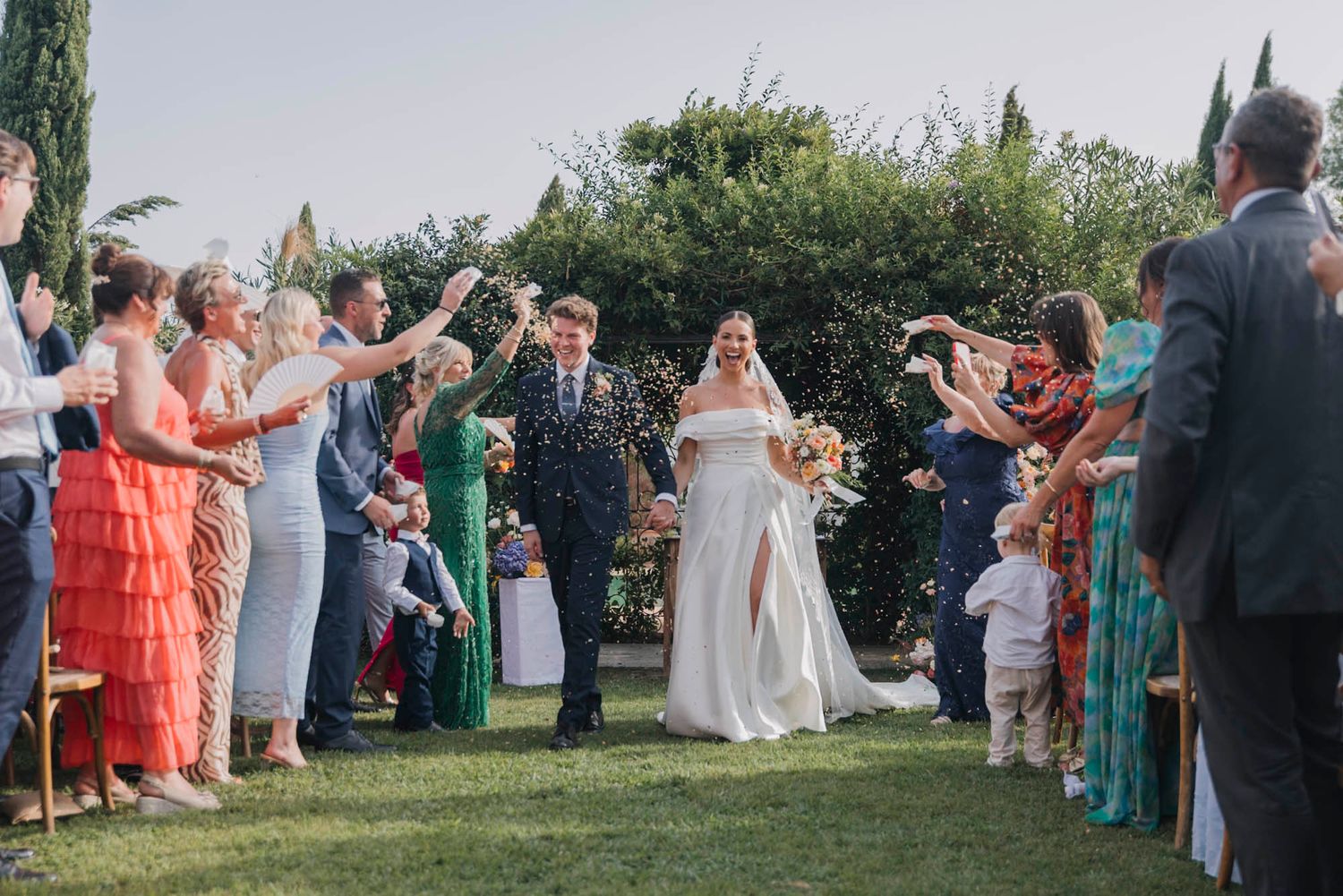 Wedding guests throwing confetti at the newlyweds during outdoor celebration.