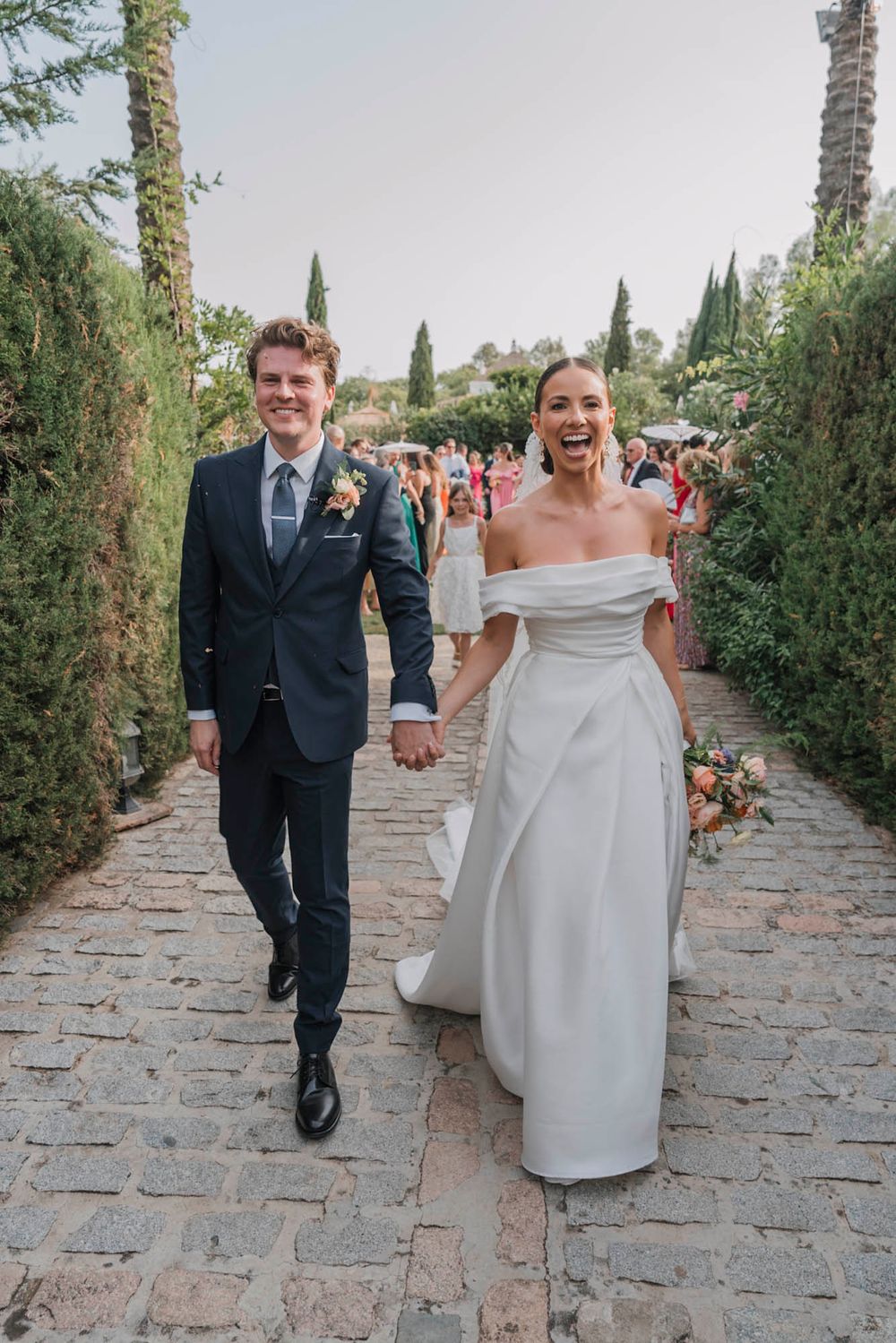 Joyful newlyweds walking down stone pathway lined with cypress trees at Tuscan venue.