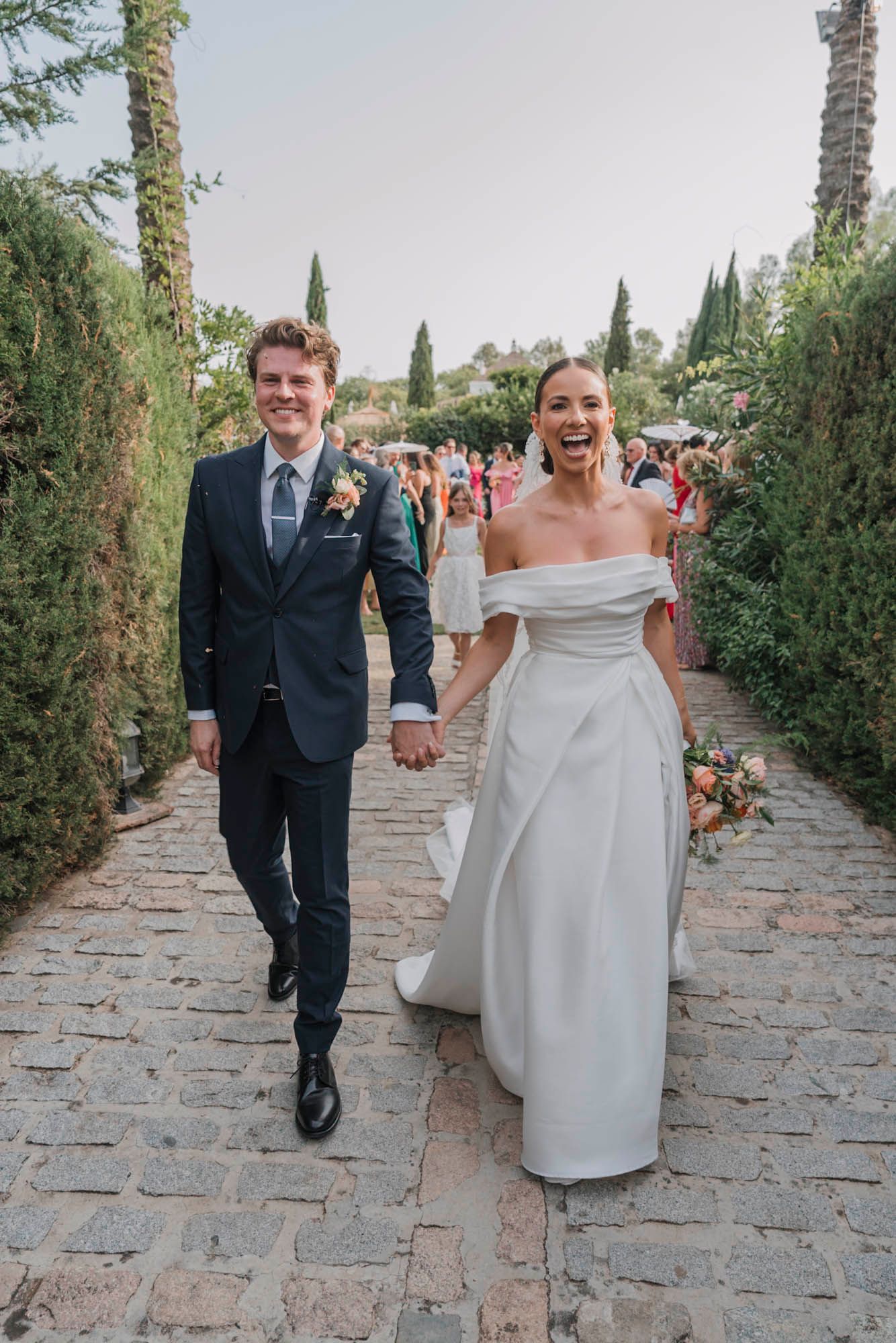 Joyful newlyweds walking down stone pathway lined with cypress trees at Tuscan venue.