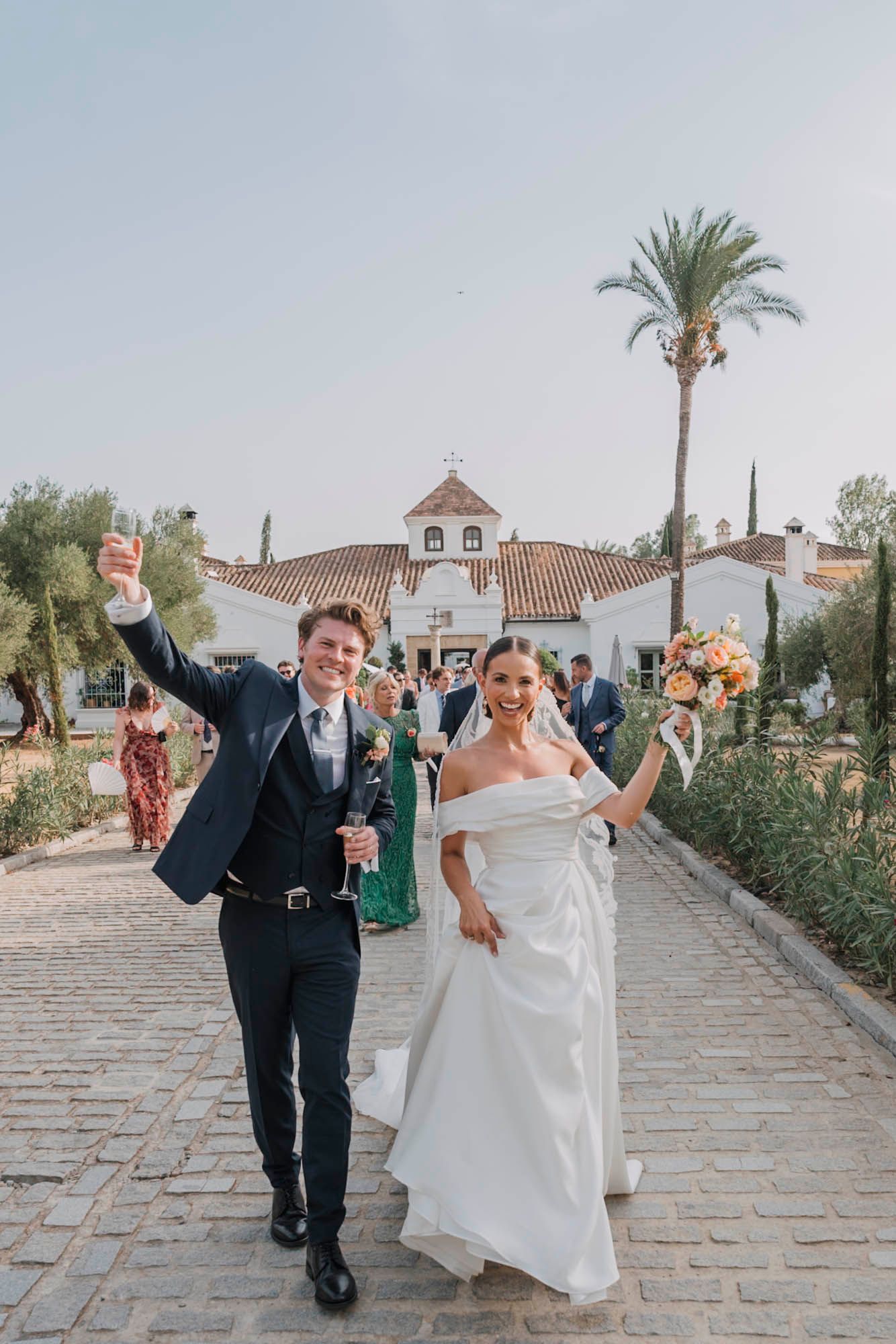 A jubilant newlywed couple walks down a stone path with a Spanish-style church and tall palm tree in the background.