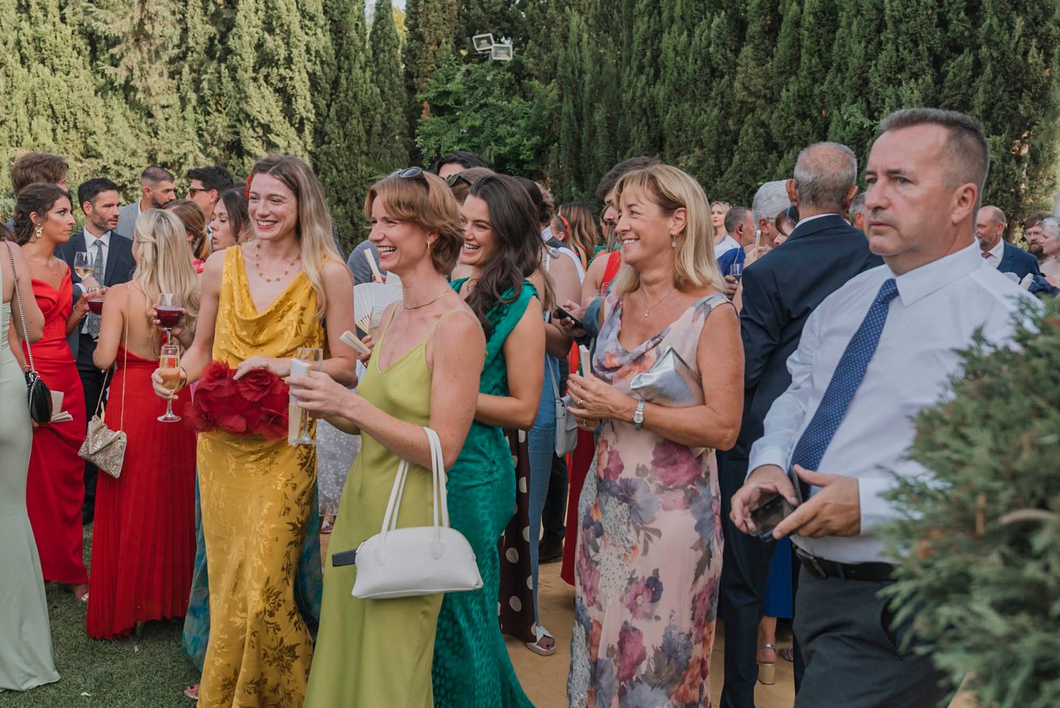 Wedding guests in colorful formal attire gather outdoors during a celebration.