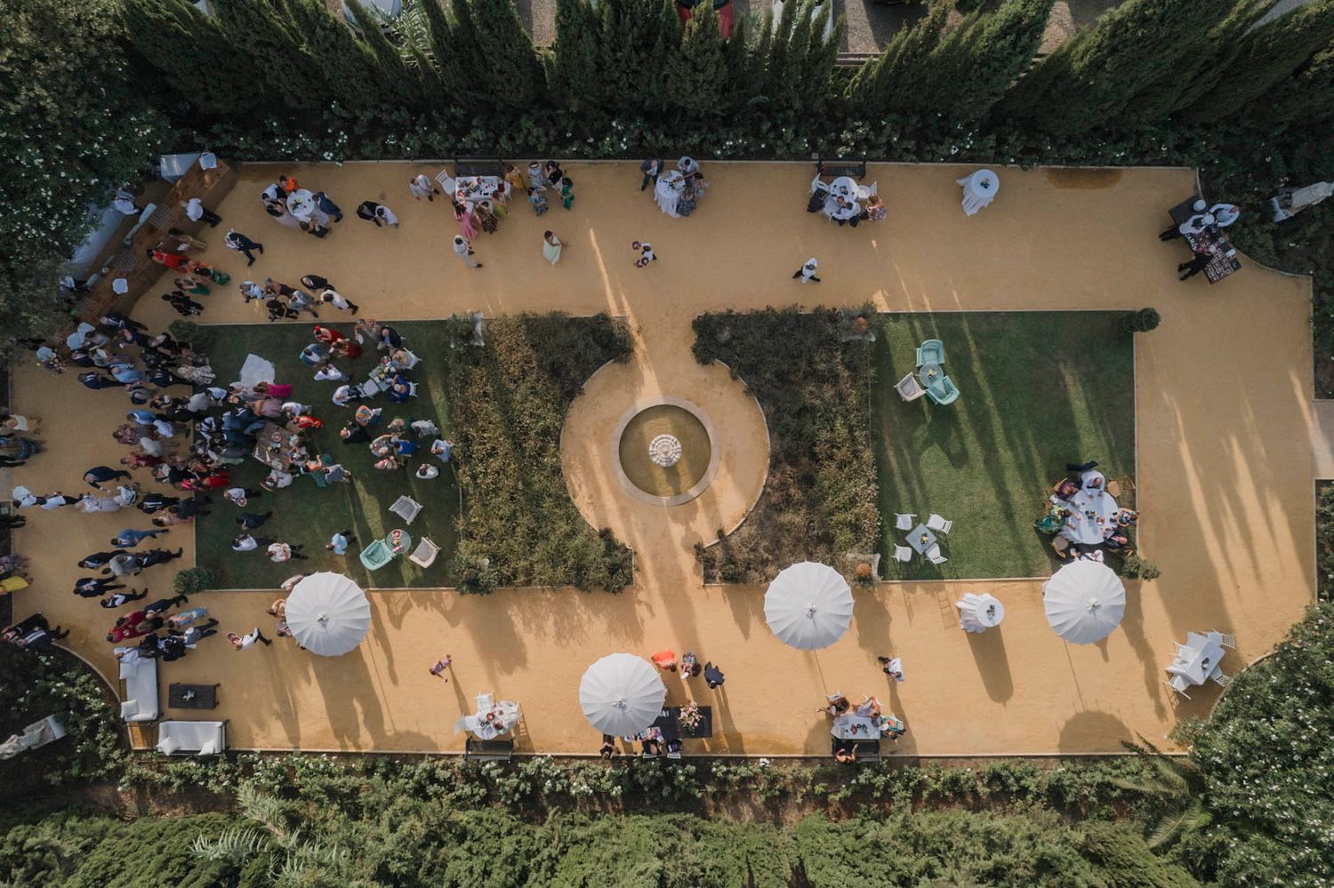 Aerial view of an outdoor wedding reception with guests seated at round tables among cypress trees.