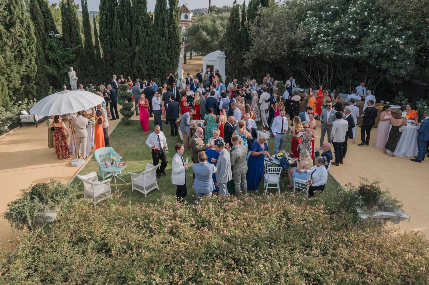 Bird's eye view of wedding guests mingling in a Mediterranean garden setting with white umbrellas.