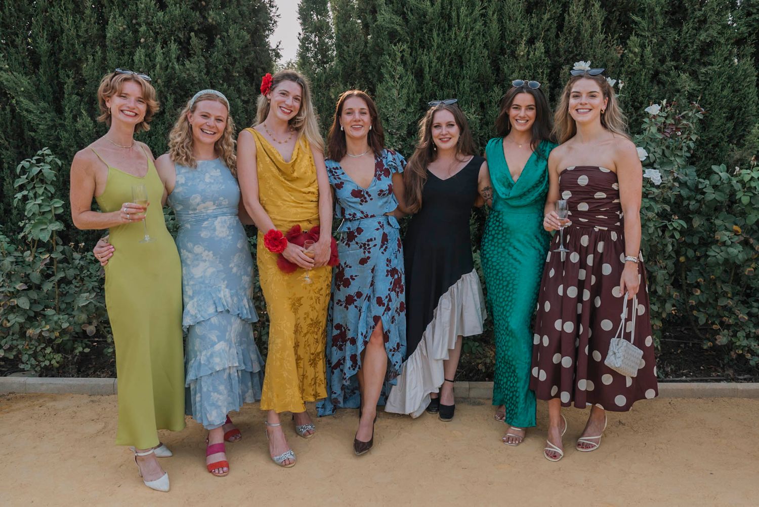 Group photo of wedding guests in vibrant formal dresses against a cypress hedge backdrop.