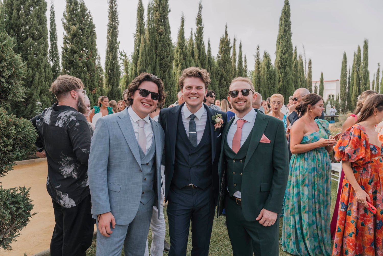 Wedding guests in suits and formal attire pose among cypress trees.