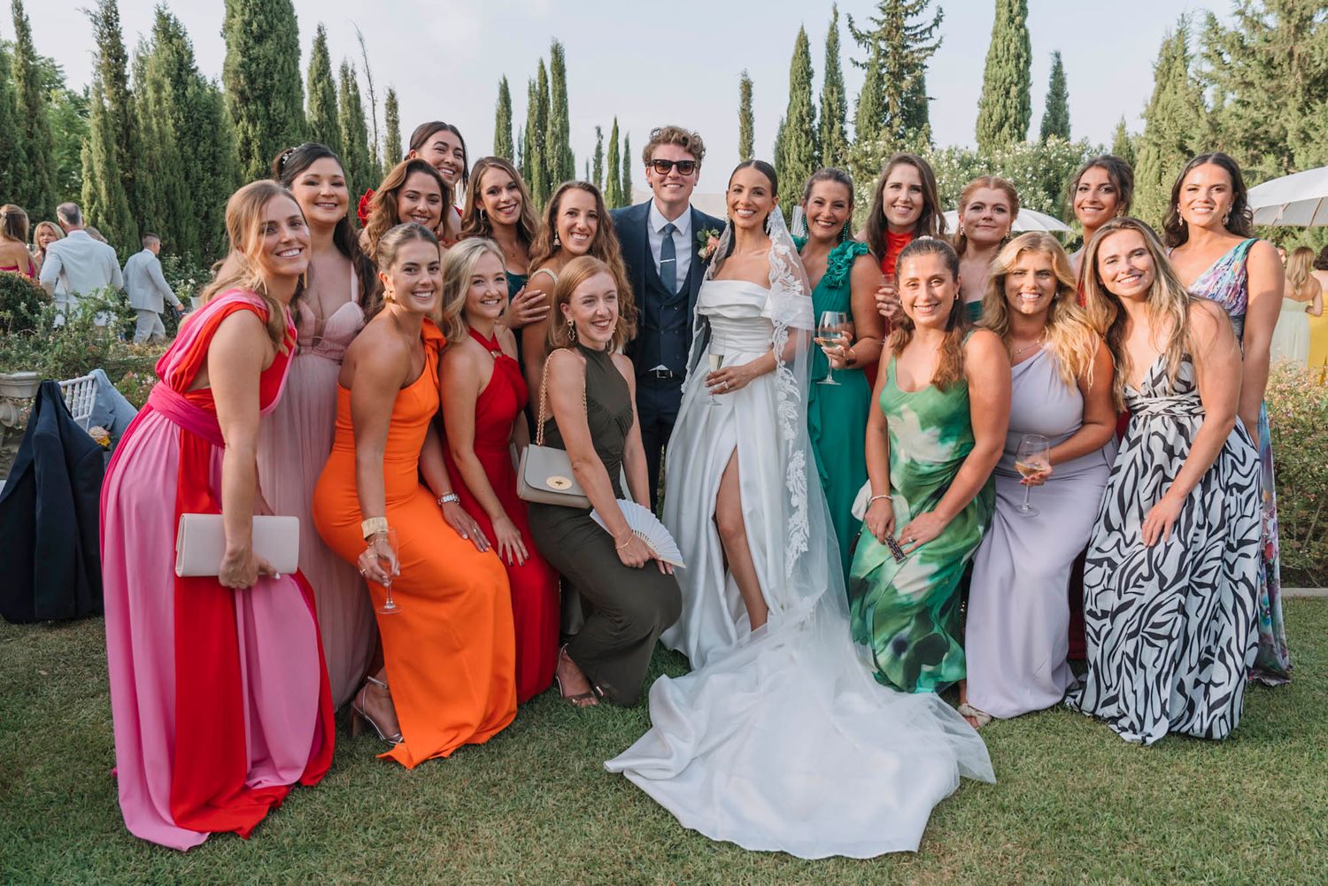 Wedding party group photo with colorful bridesmaid dresses in a garden setting with cypress trees in background.