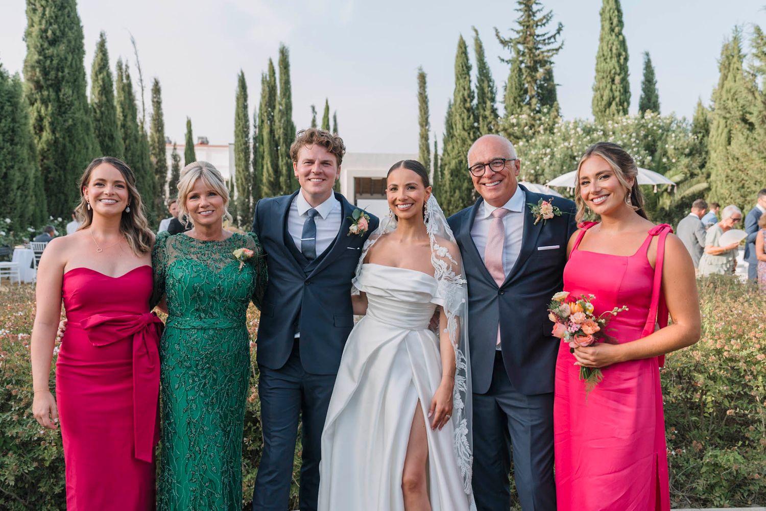 Wedding party members in bright formal attire pose together against backdrop of tall cypress trees.