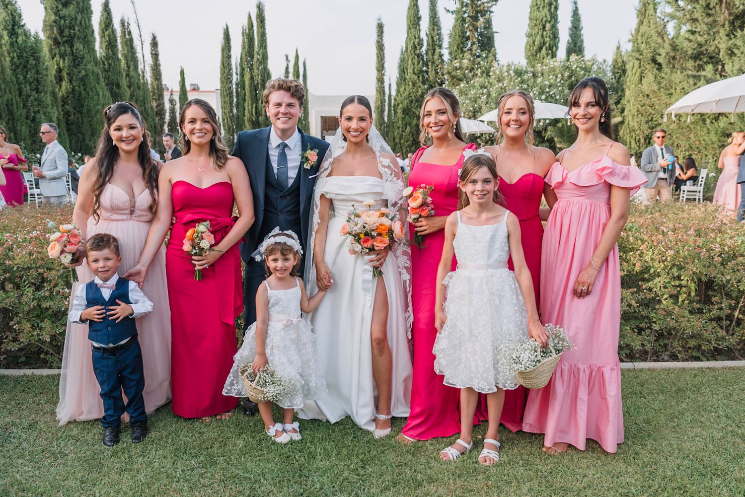 Wedding party photo featuring bridesmaids in pink dresses and flower girls in white on a garden lawn.