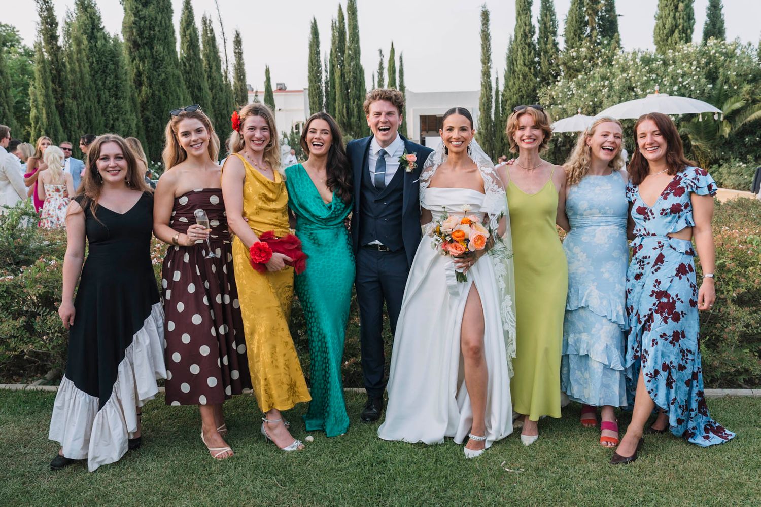 Bridal party wearing colorful mix-and-match dresses poses together on garden grounds.