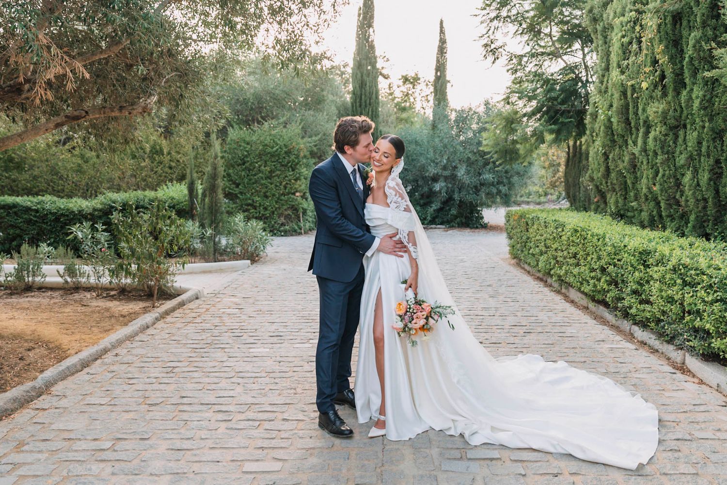 Romantic wedding portrait of couple embracing on stone path surrounded by cypress trees.