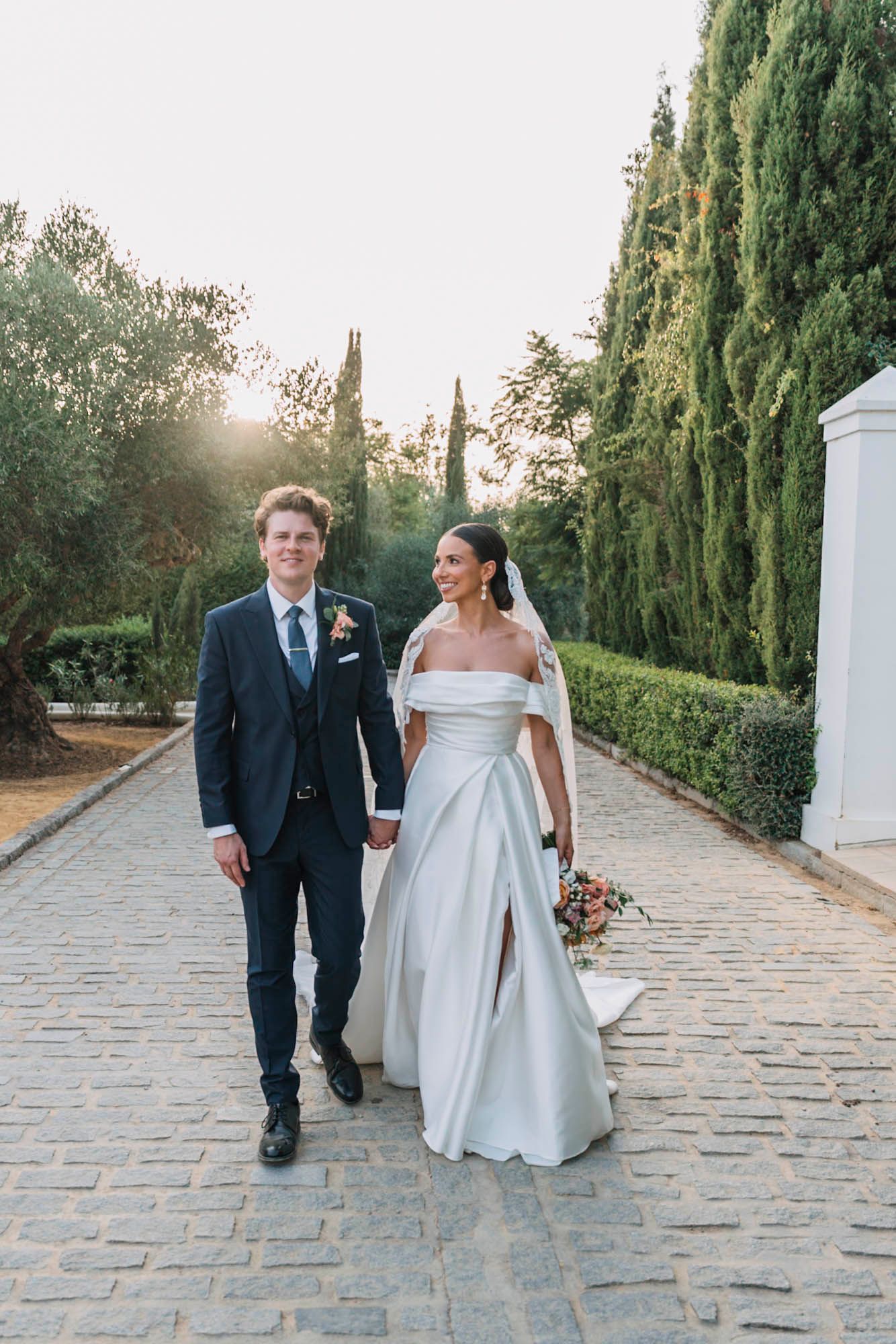 Elegant couple strolling on cobblestone path with cypress trees and garden backdrop.