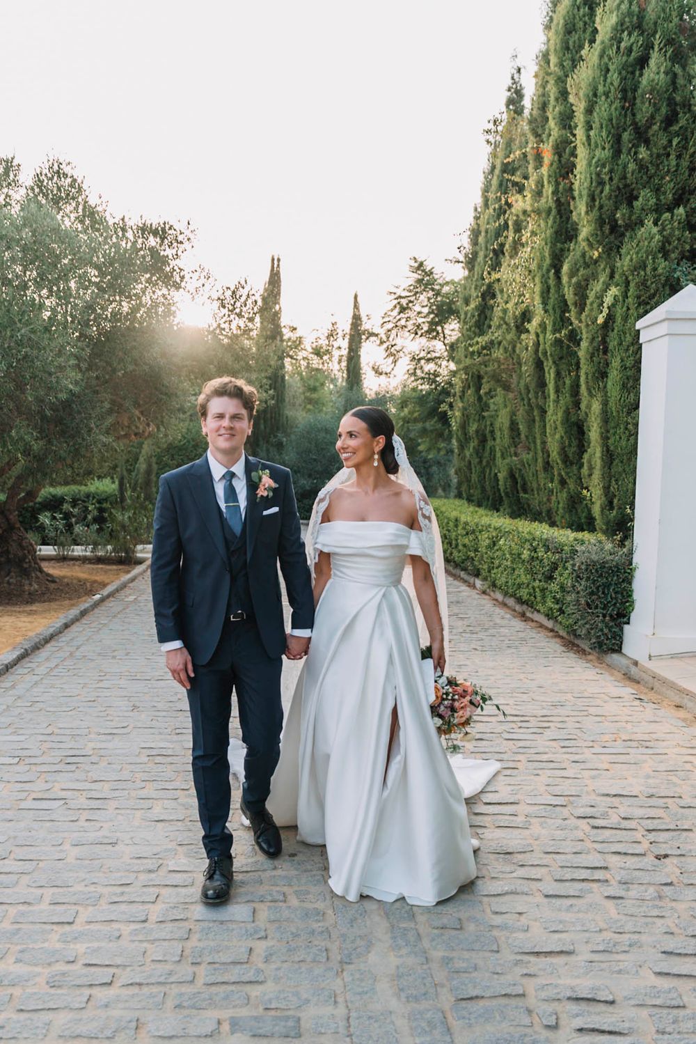 Elegant couple strolling on cobblestone path with cypress trees and garden backdrop.