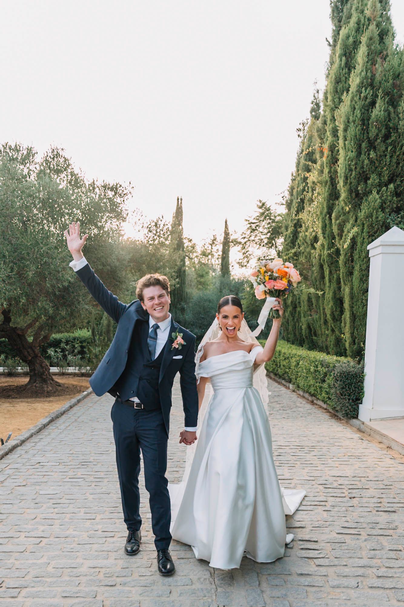 Newlyweds celebrate joyfully on a stone path lined with cypress trees and olive groves.