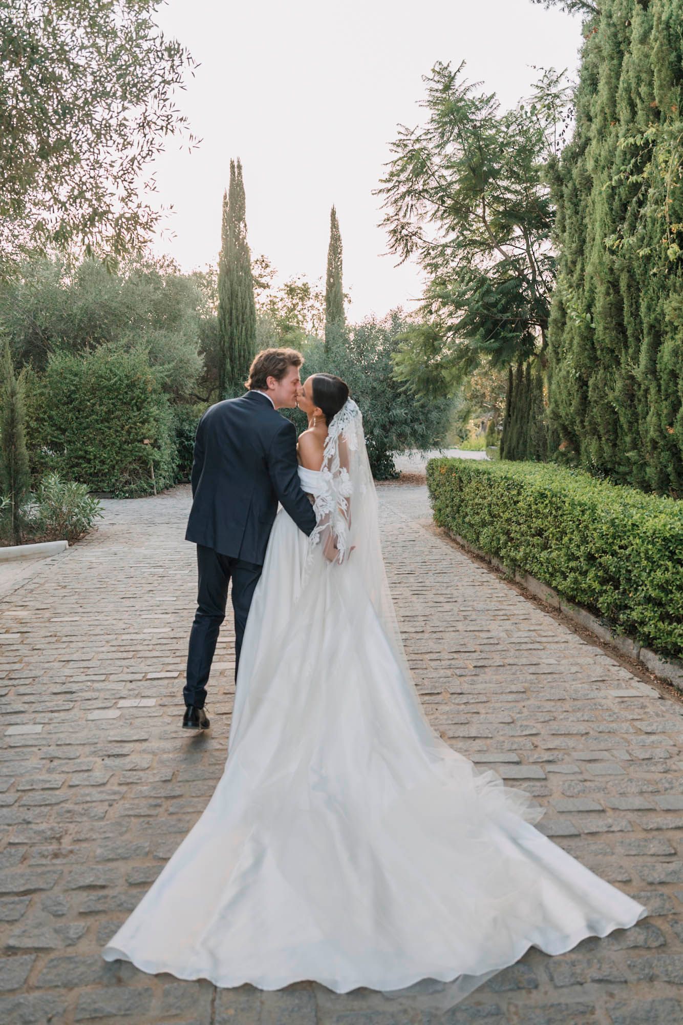Elegant wedding couple shares an intimate moment on a cobblestone path surrounded by greenery.