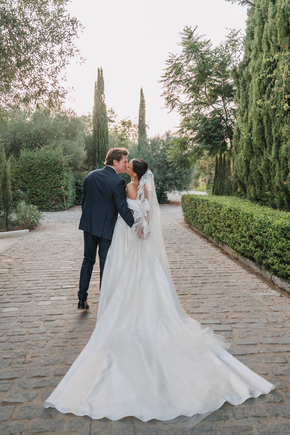 Elegant wedding couple shares an intimate moment on a cobblestone path surrounded by greenery.