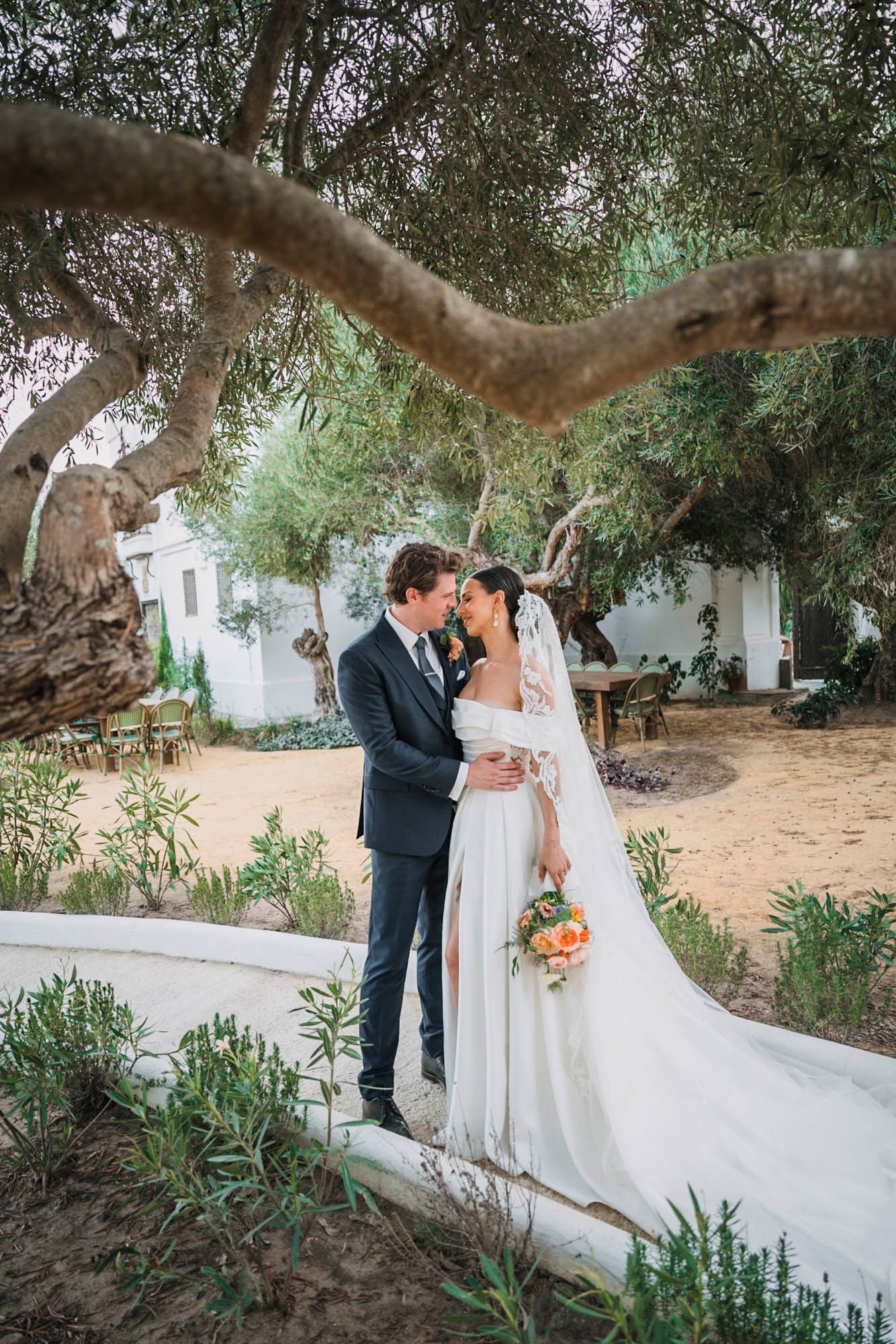 Wedding couple embraces under a twisted olive tree branch in a Mediterranean garden setting.