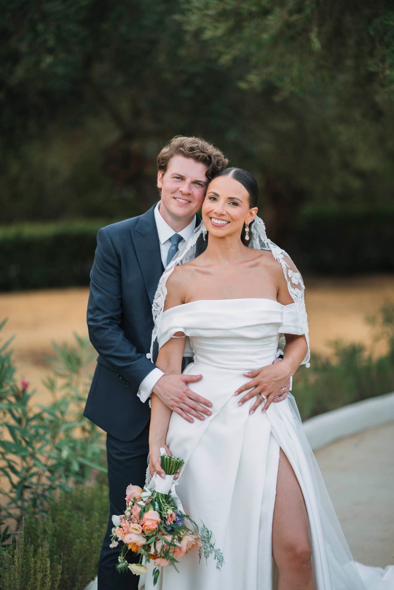Wedding couple poses in garden setting with bride holding peach and coral bouquet.