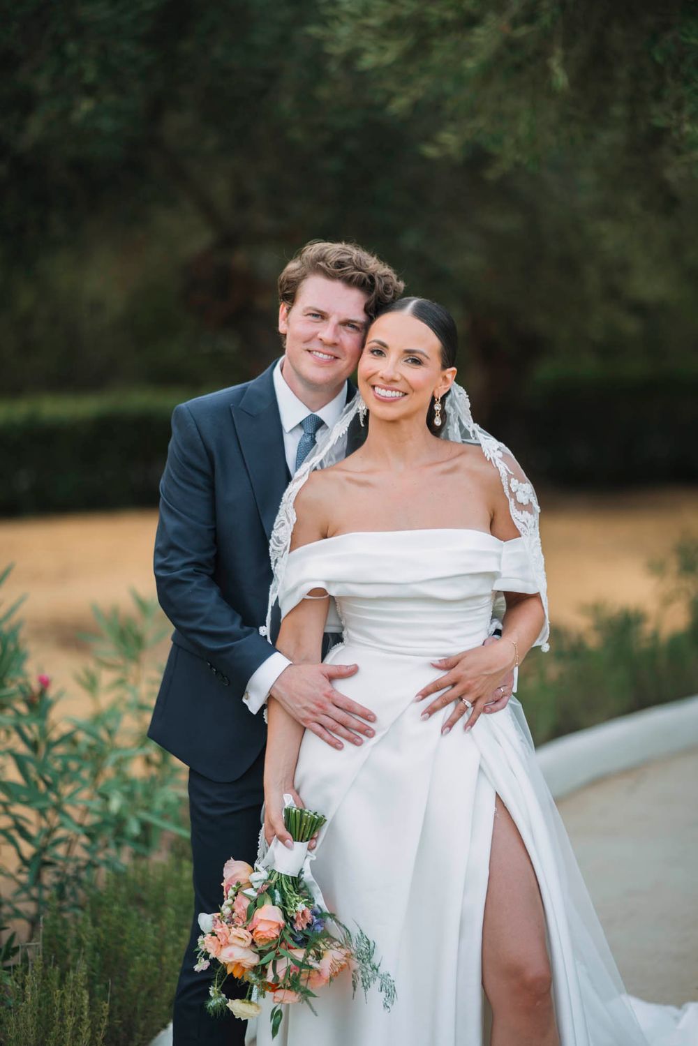 Wedding couple poses in garden setting with bride holding peach and coral bouquet.