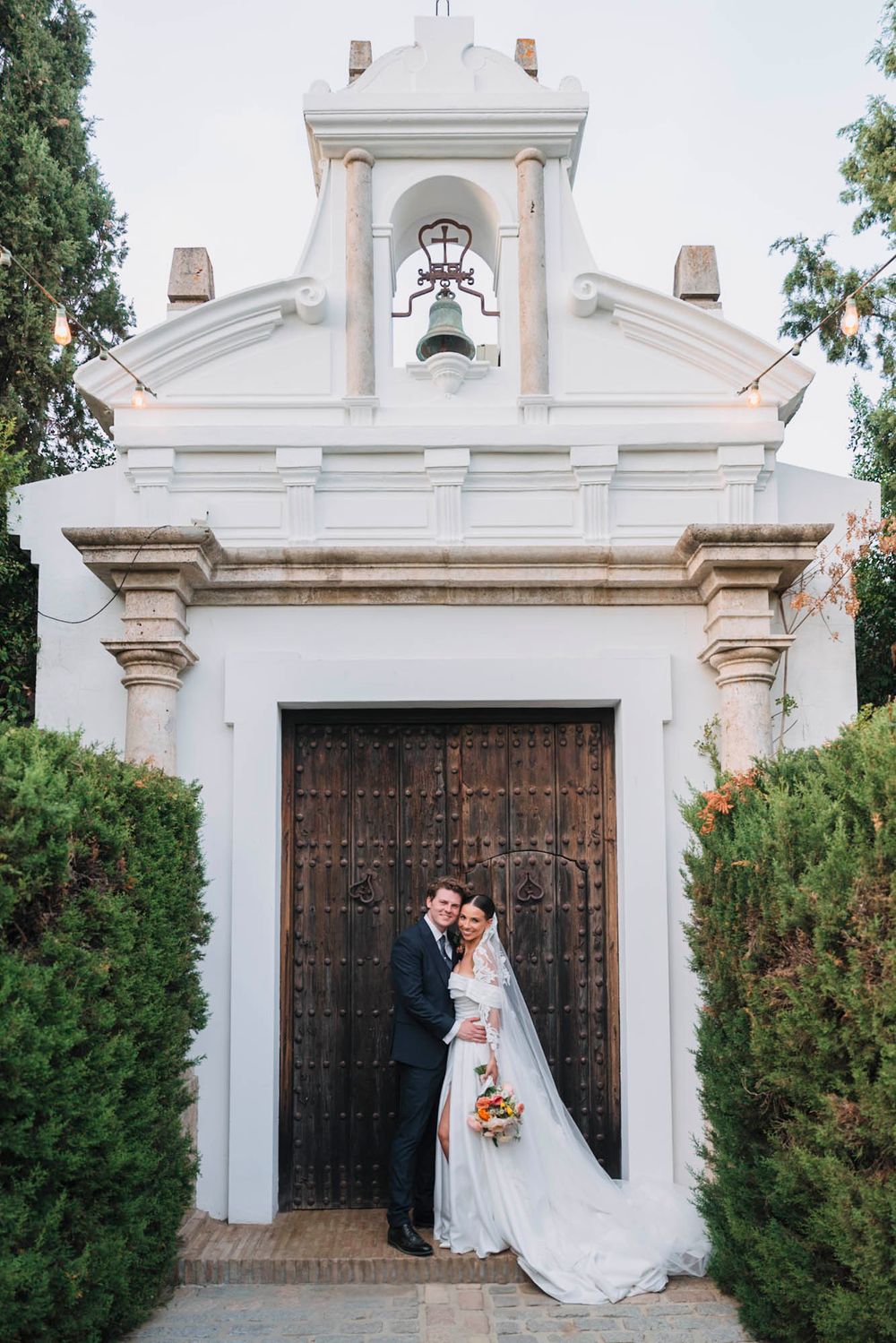 Wedding couple poses at white Spanish chapel entrance with ornate wooden doors.
