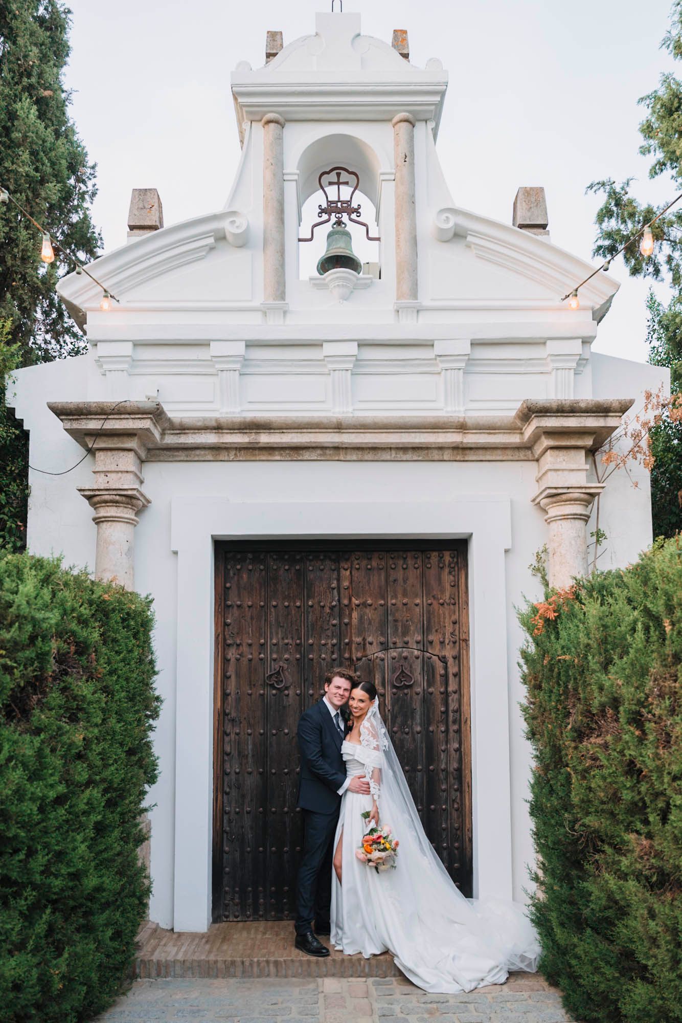 Wedding couple poses at white Spanish chapel entrance with ornate wooden doors.