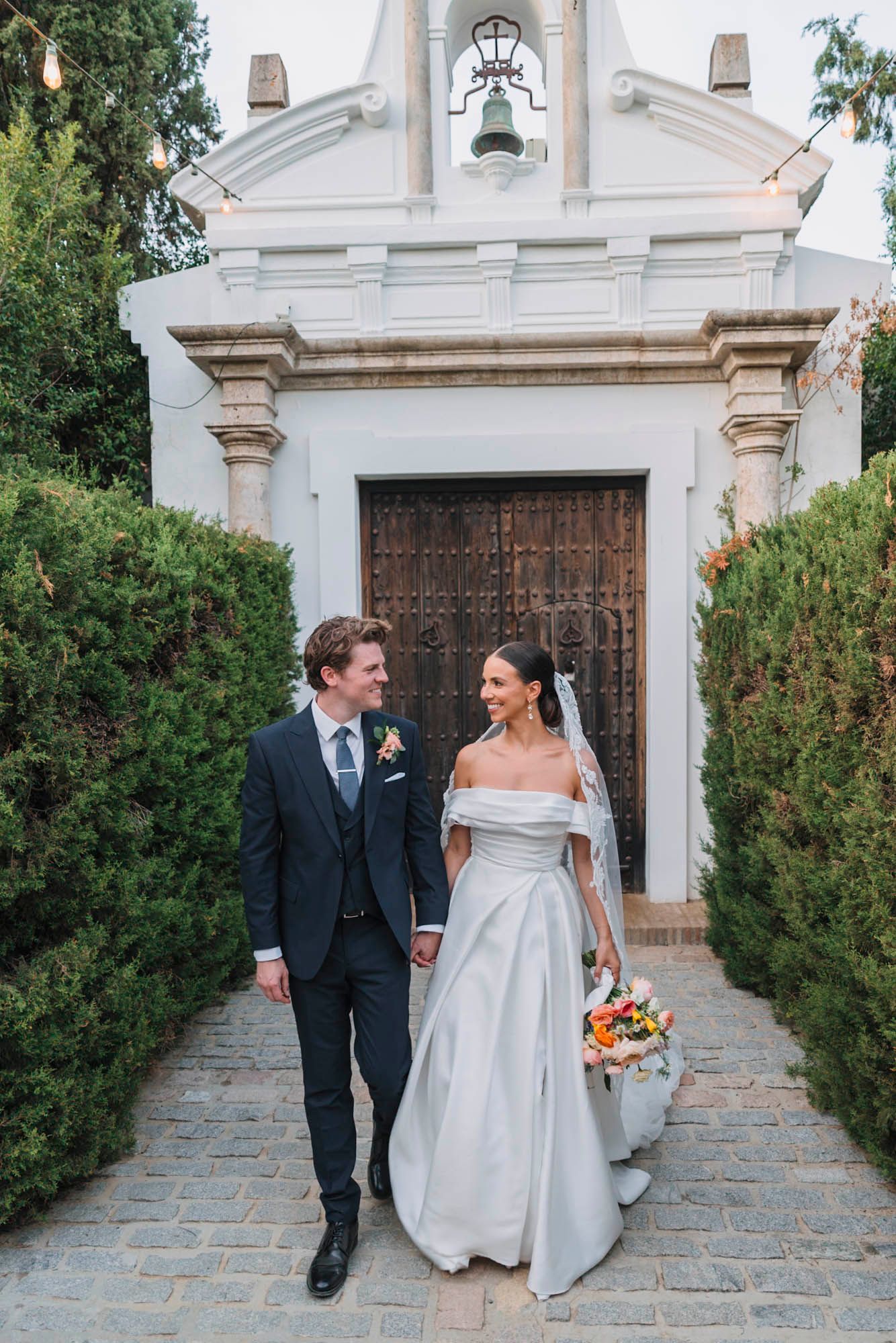 Newlyweds walk along pathway in front of white Spanish chapel with bell tower.