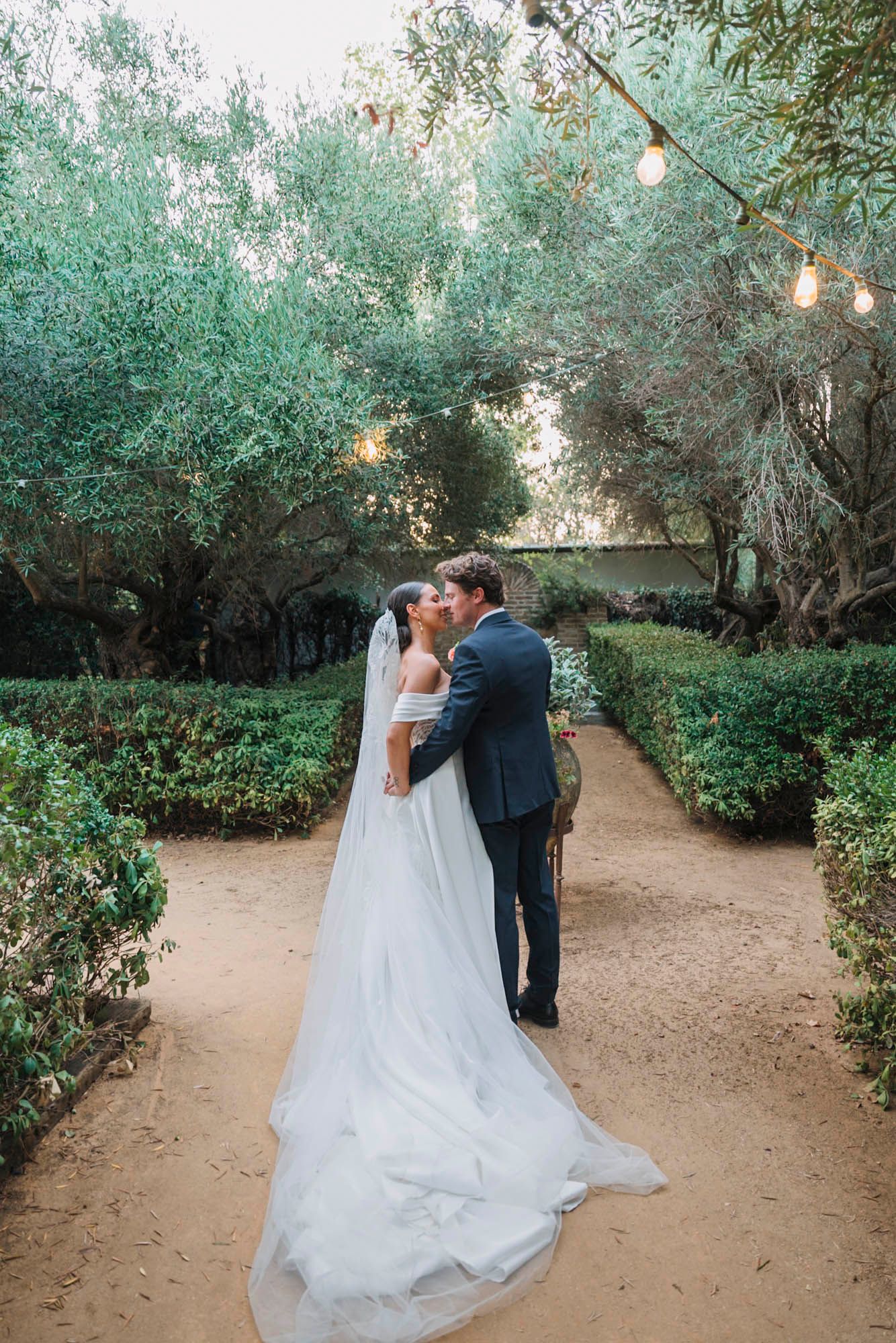 Wedding couple shares romantic moment under string lights in garden path.