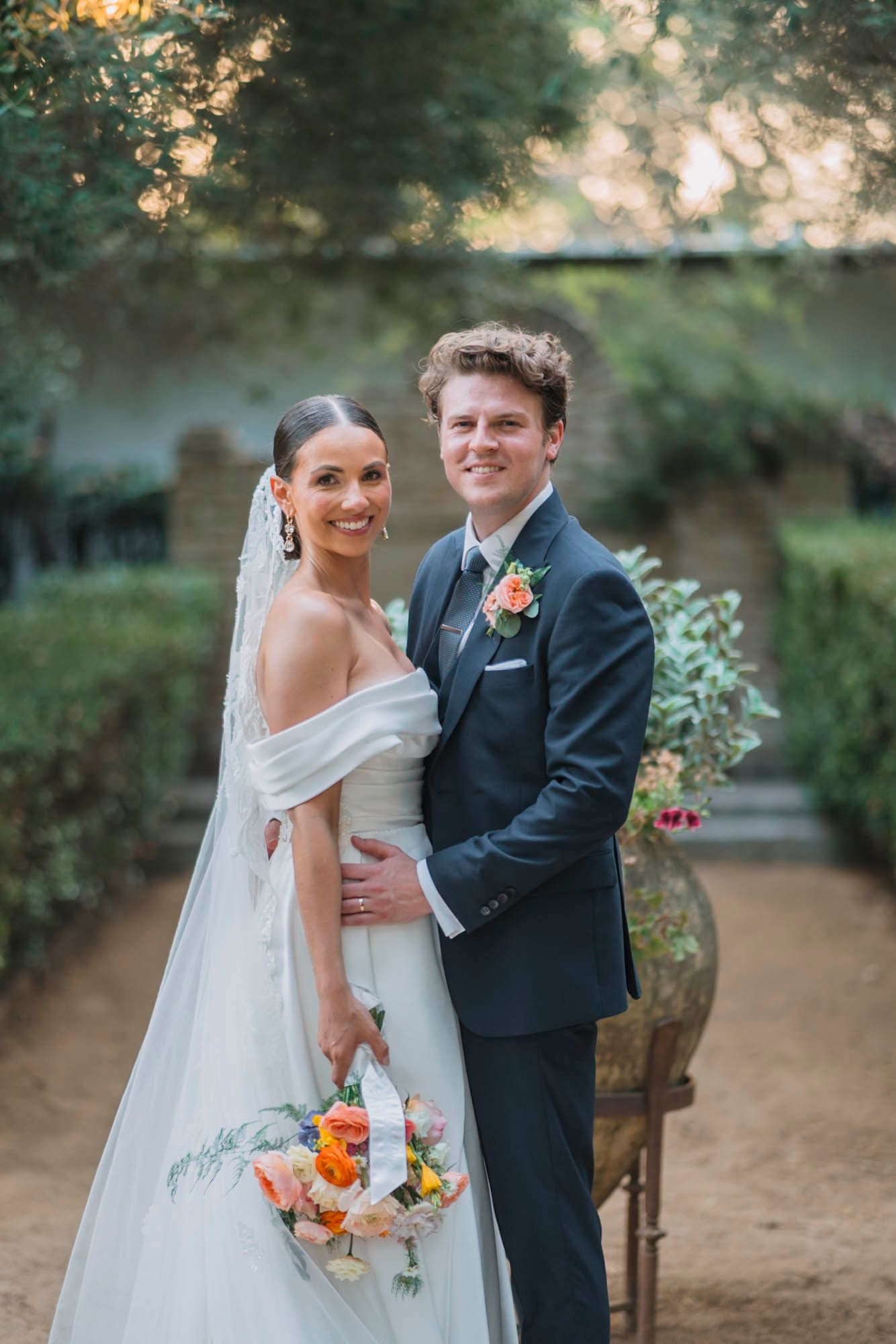 Elegant wedding portrait in lush garden with bride holding coral floral bouquet.