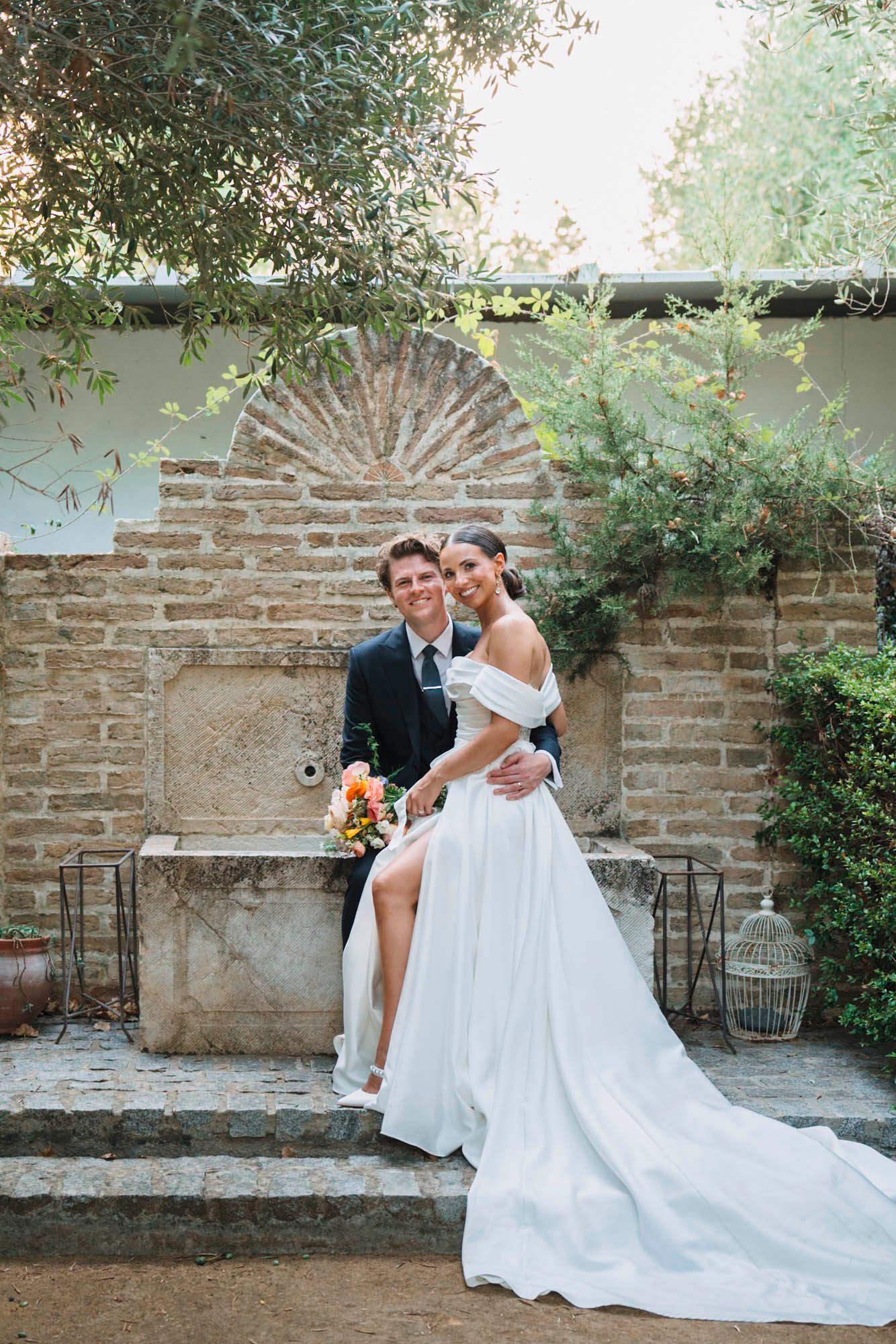 Wedding couple poses on stone bench against rustic Mediterranean wall.