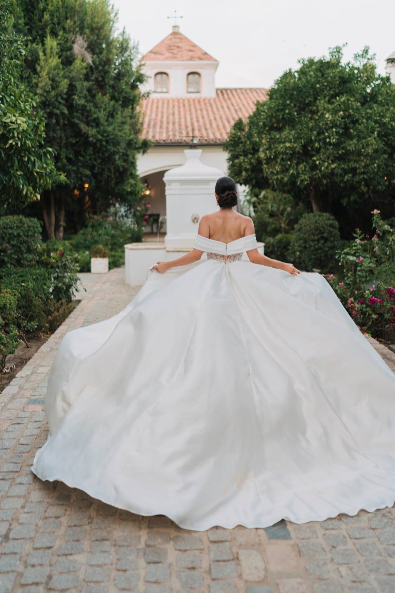 A stunning white wedding gown billows dramatically against a Spanish mission-style church backdrop with terracotta roof tiles.