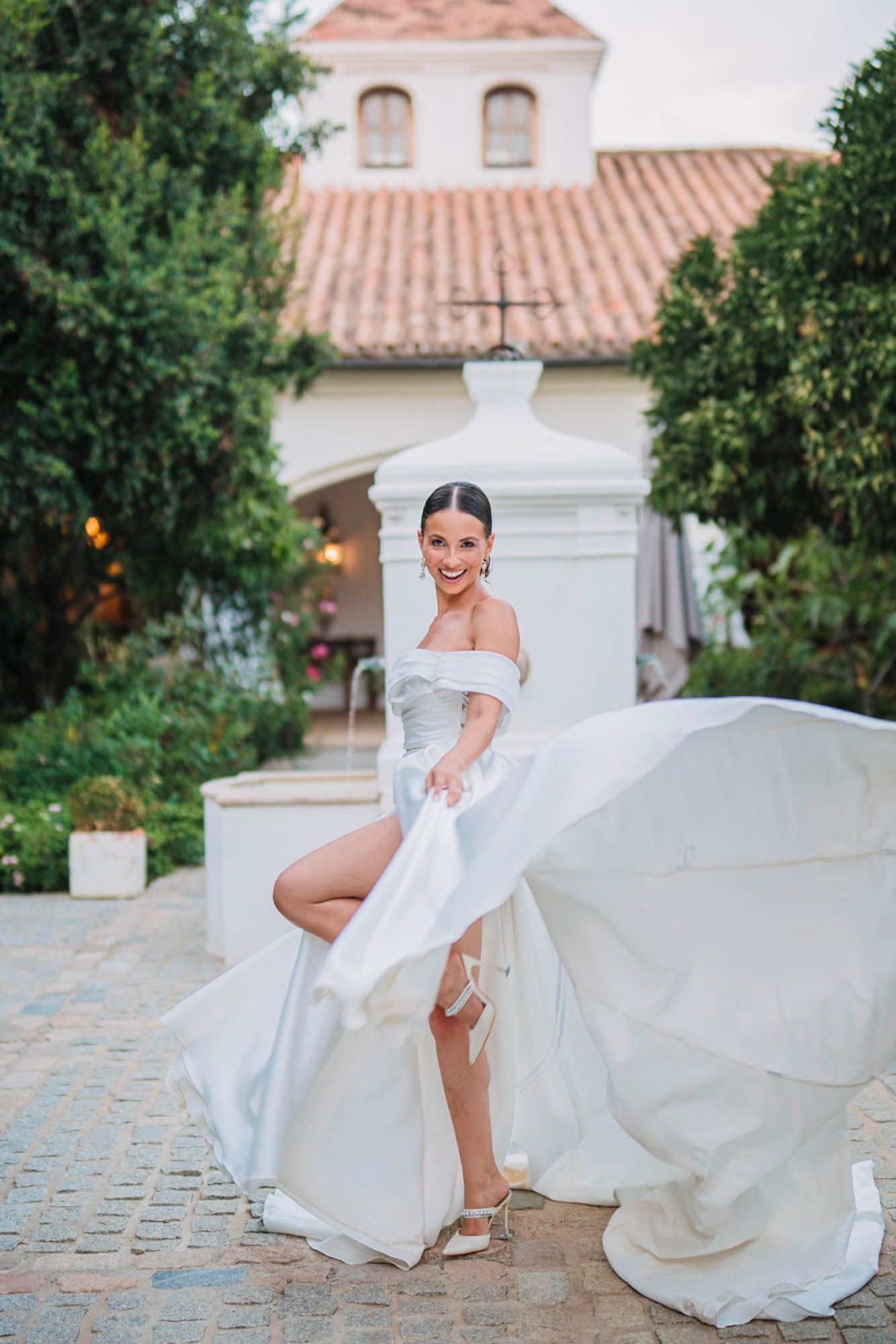 An elegant off-shoulder wedding dress flows gracefully on a cobblestone path in front of a historic church.