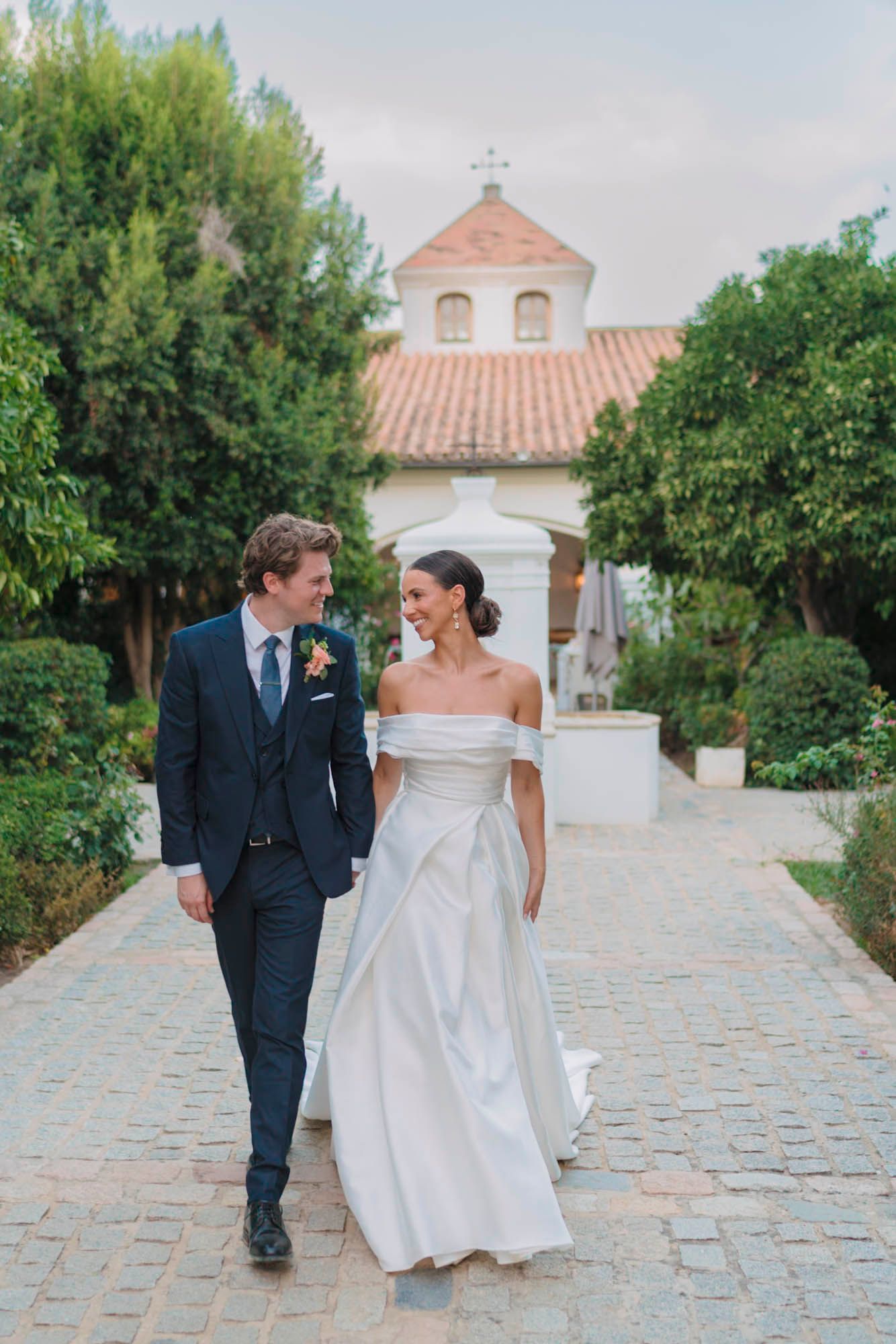 A wedding couple walks along a cobblestone path leading to a Spanish mission-style church.