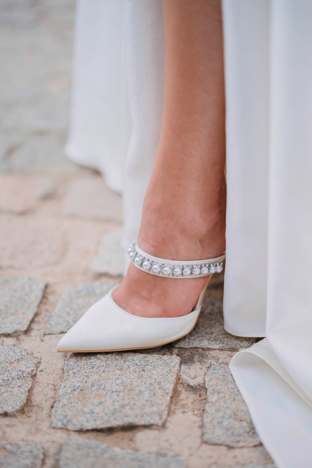 An elegant white bridal shoe with crystal embellishments rests on cobblestone pavement.