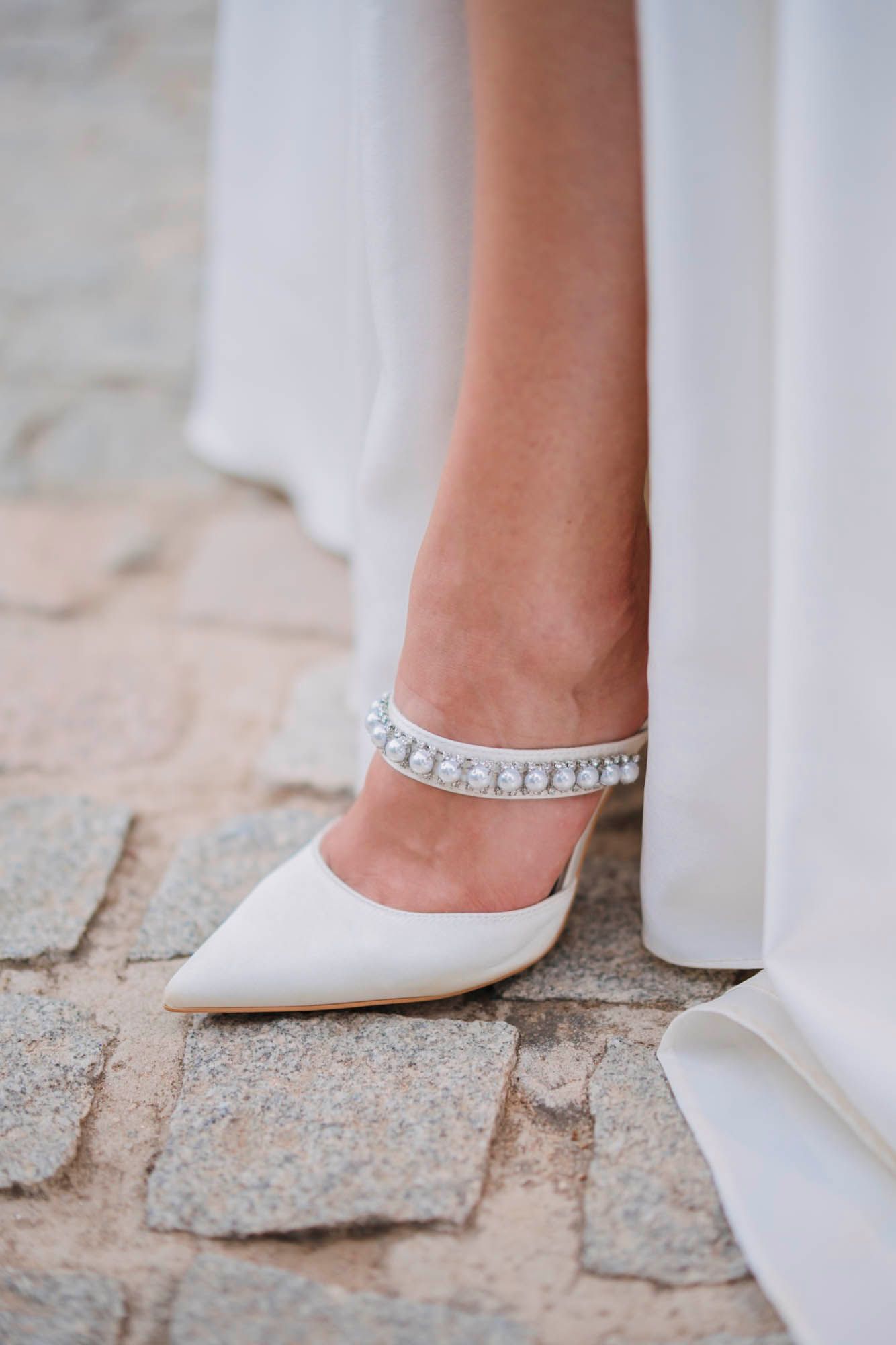 An elegant white bridal shoe with crystal embellishments rests on cobblestone pavement.