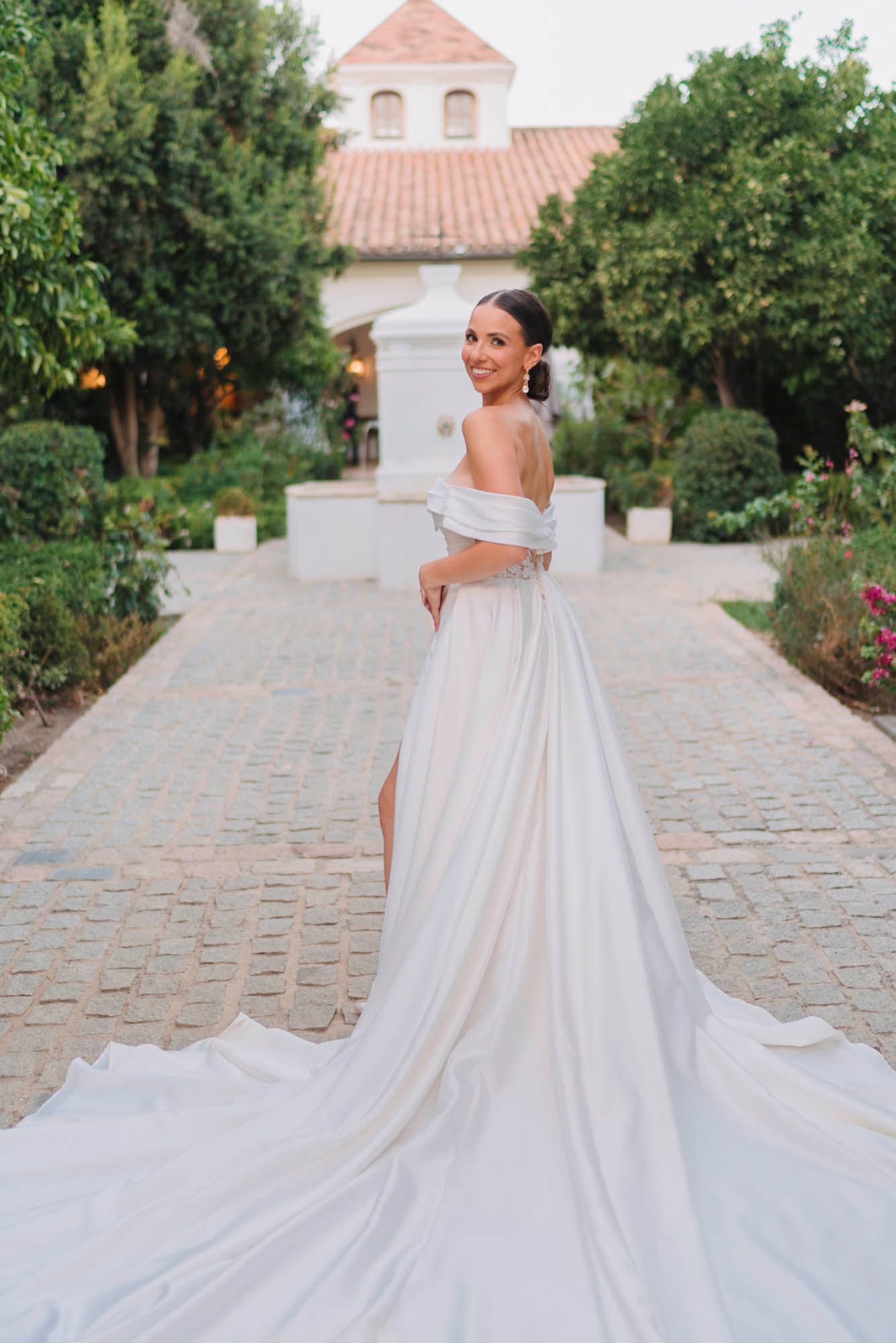 Bride in flowing off-shoulder gown poses in garden courtyard with white chapel in background.