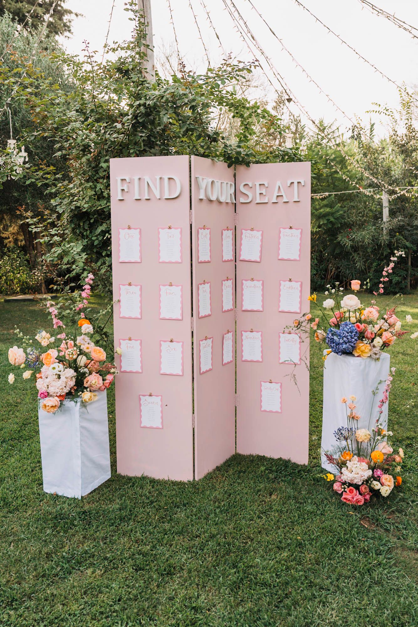 Pink find-your-seat display board decorated with colorful floral arrangements for wedding reception.