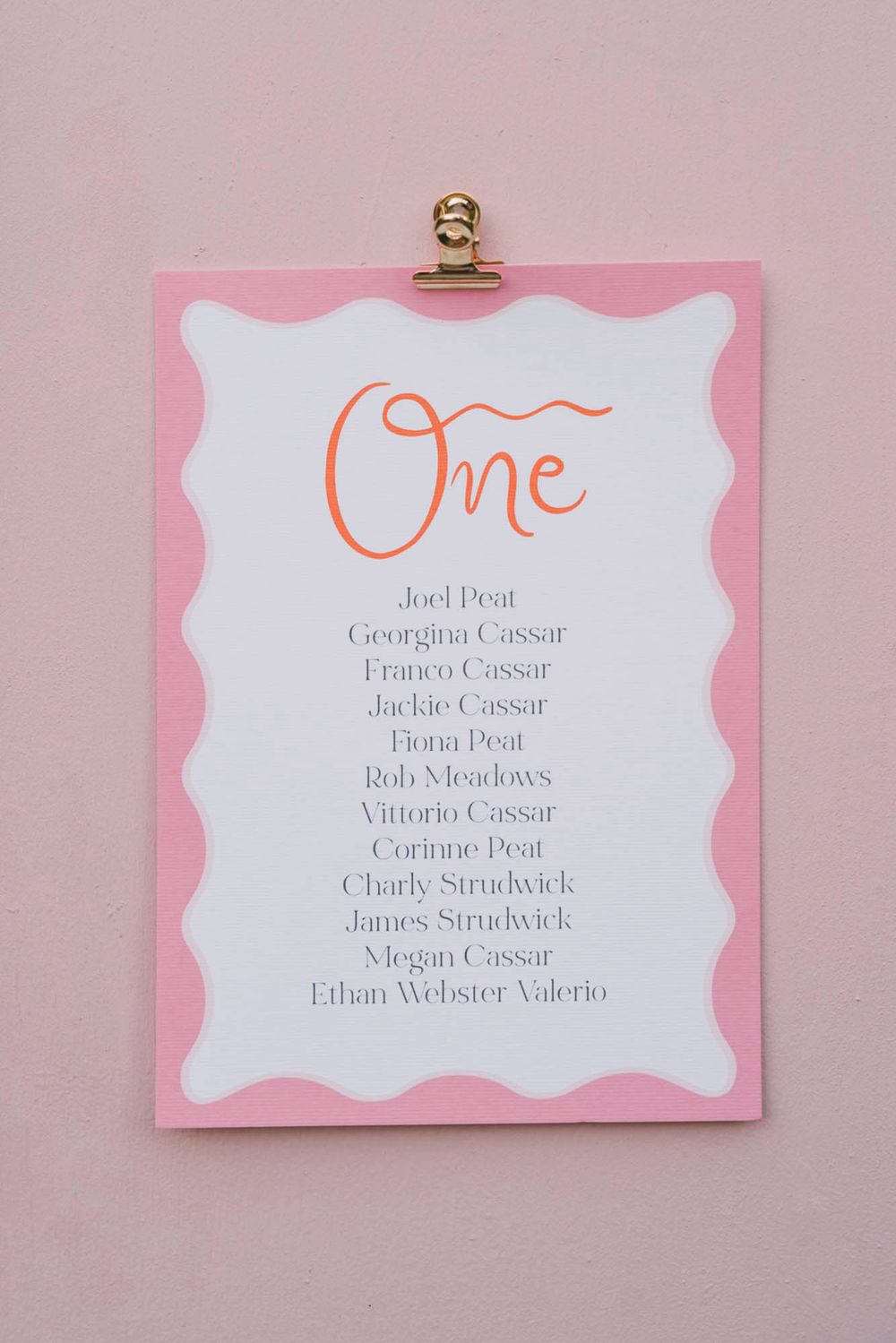 Pink and white wedding table seating chart with elegant calligraphy listing guest names in orange script.