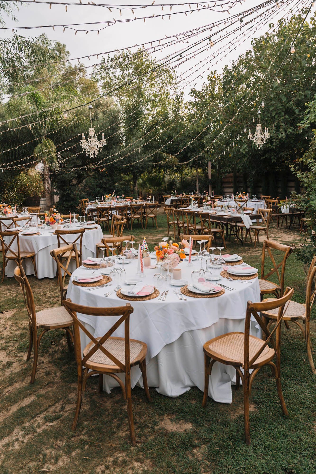 Elegant outdoor wedding reception setup with round tables, white linens and wooden crossback chairs.