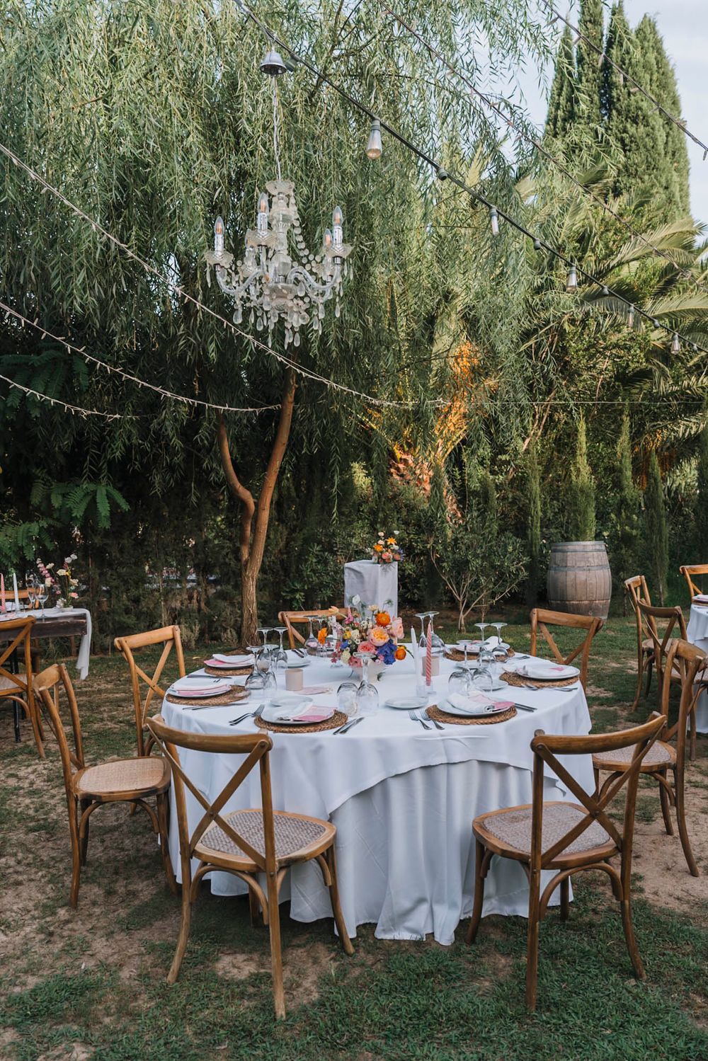 Romantic garden wedding reception table with crystal chandelier and white linens under willow trees.