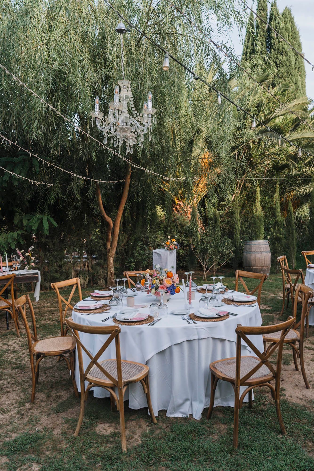 Romantic garden wedding reception table with crystal chandelier and white linens under willow trees.
