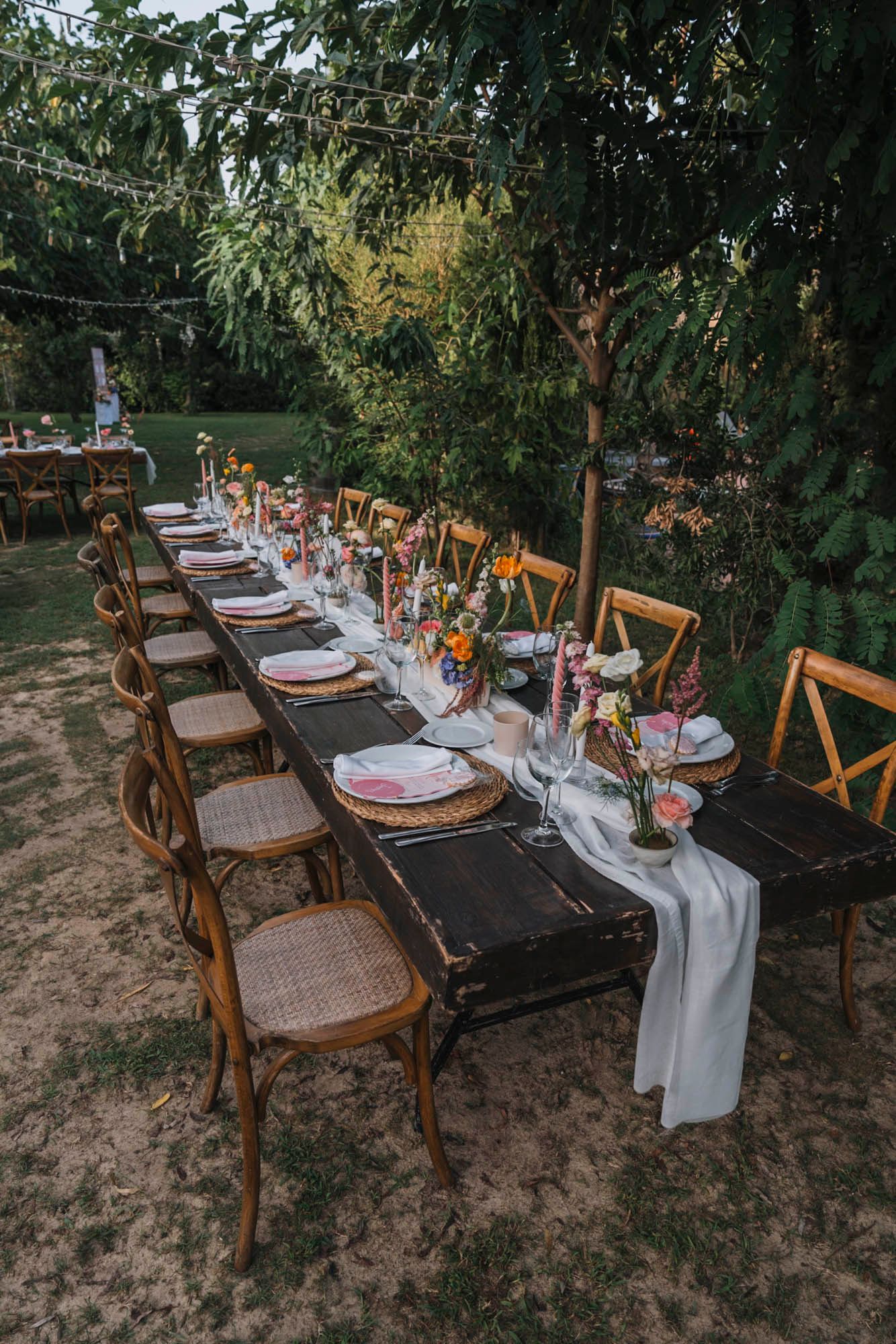 Long wooden farm table set for wedding dinner with cross-back chairs and elegant place settings.