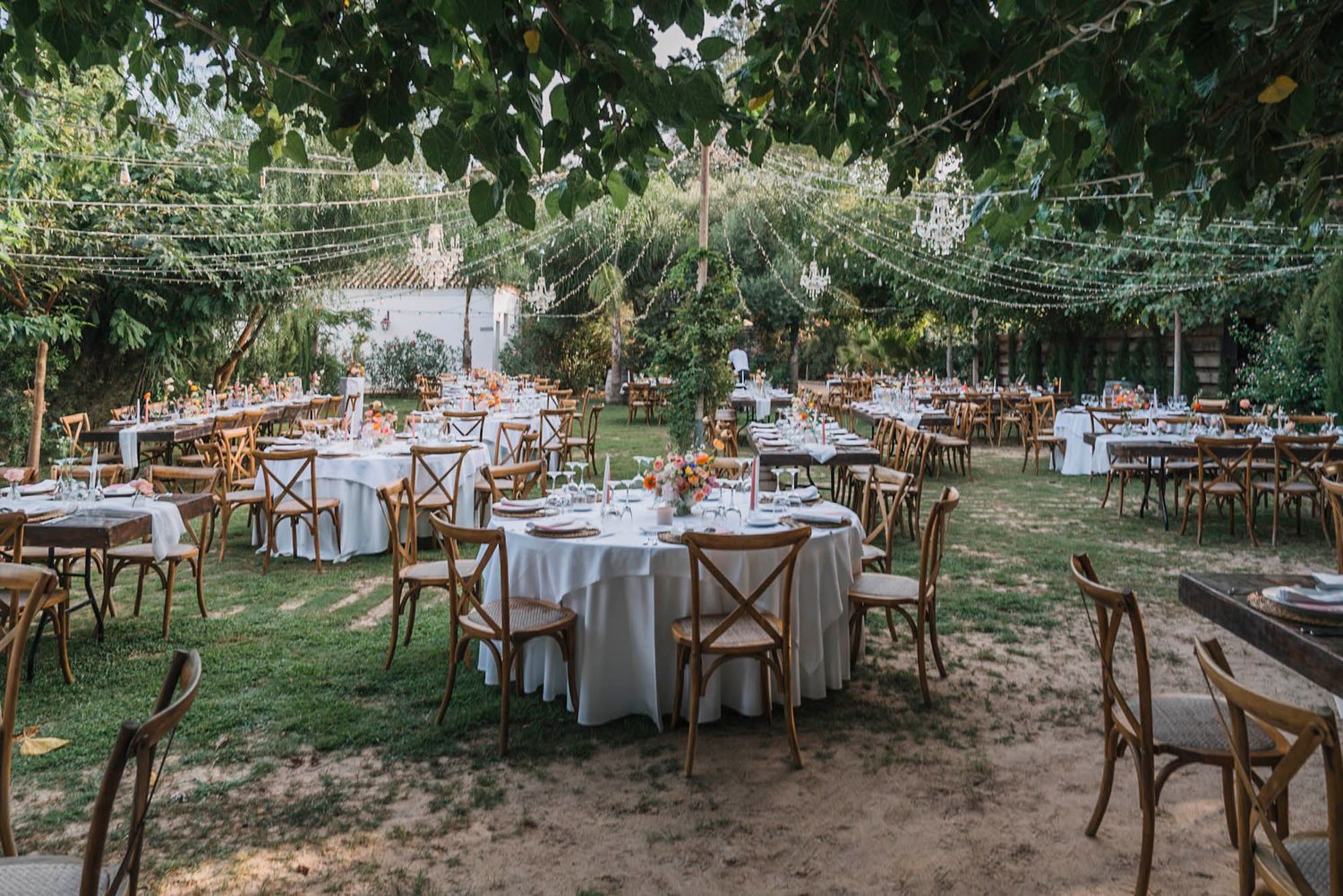 Garden wedding reception area with round tables and wooden chairs arranged under string lights.