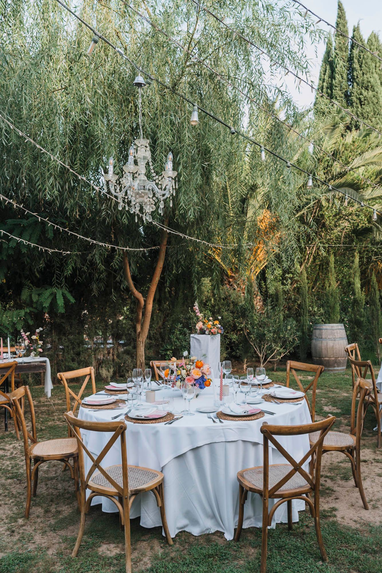 Elegant outdoor wedding table setting with crystal chandelier hanging from willow tree and rustic wooden chairs.