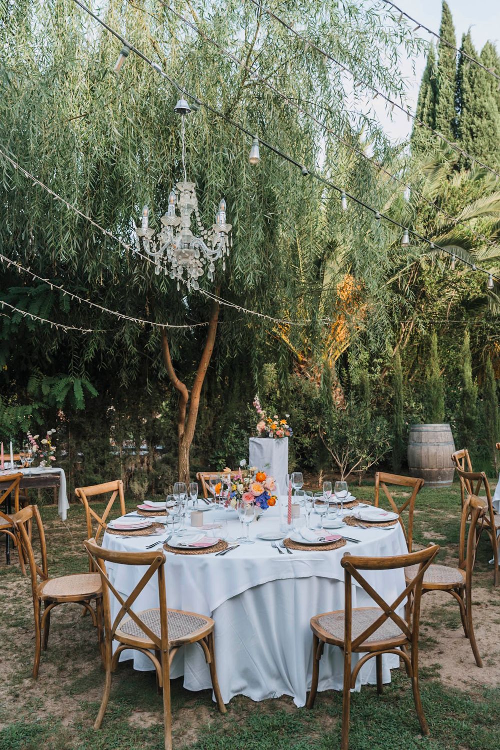 Elegant outdoor wedding table setting with crystal chandelier hanging from willow tree and rustic wooden chairs.