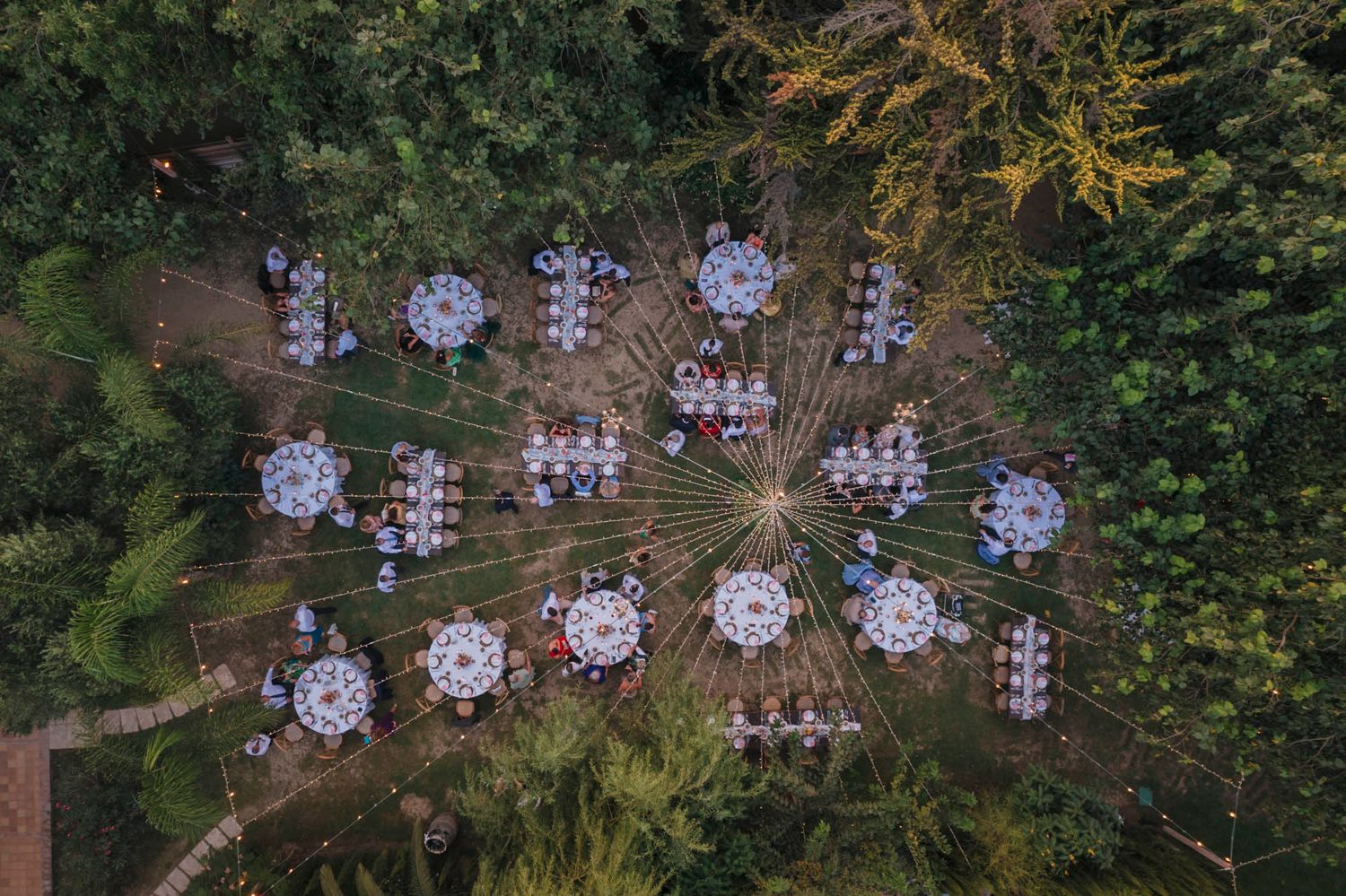 Aerial view of circular wedding reception tables arranged in garden setting with string lights overhead.