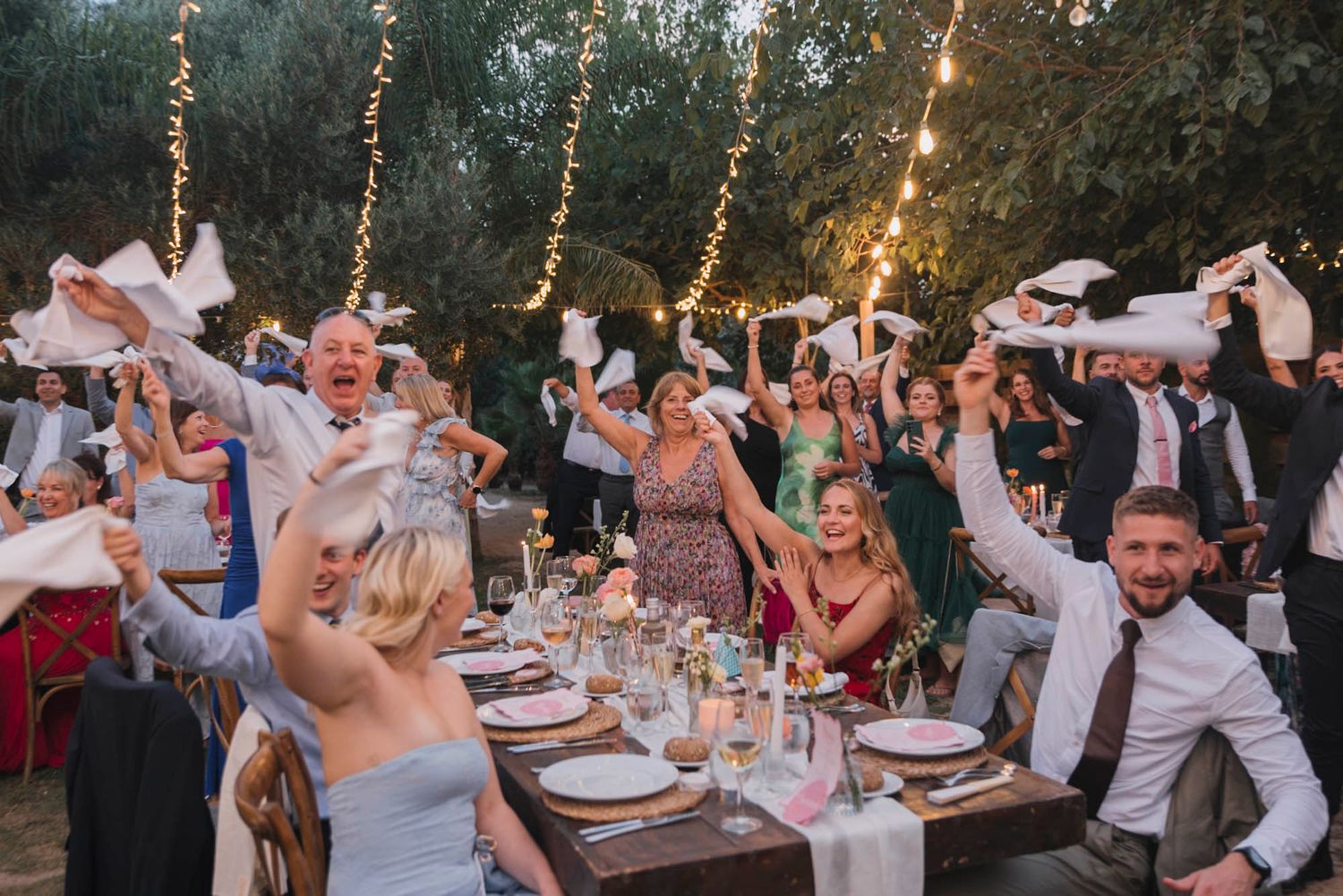 Wedding guests celebrating with raised arms at outdoor reception under string lights.