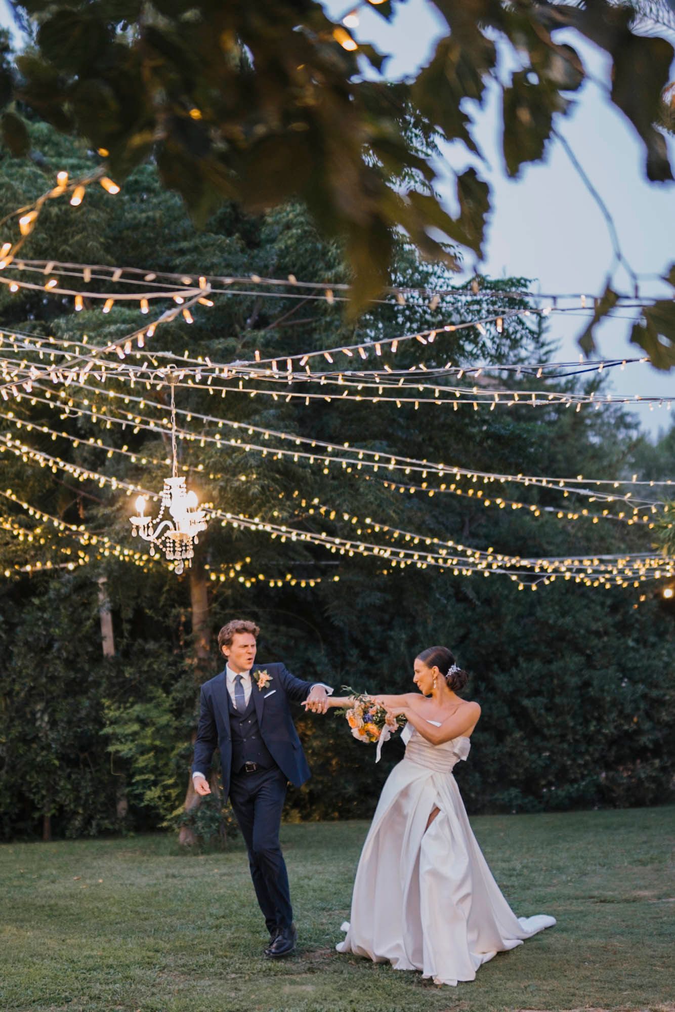 A couple plays around during their wedding reception under twinkling string lights at dusk in an outdoor garden setting.