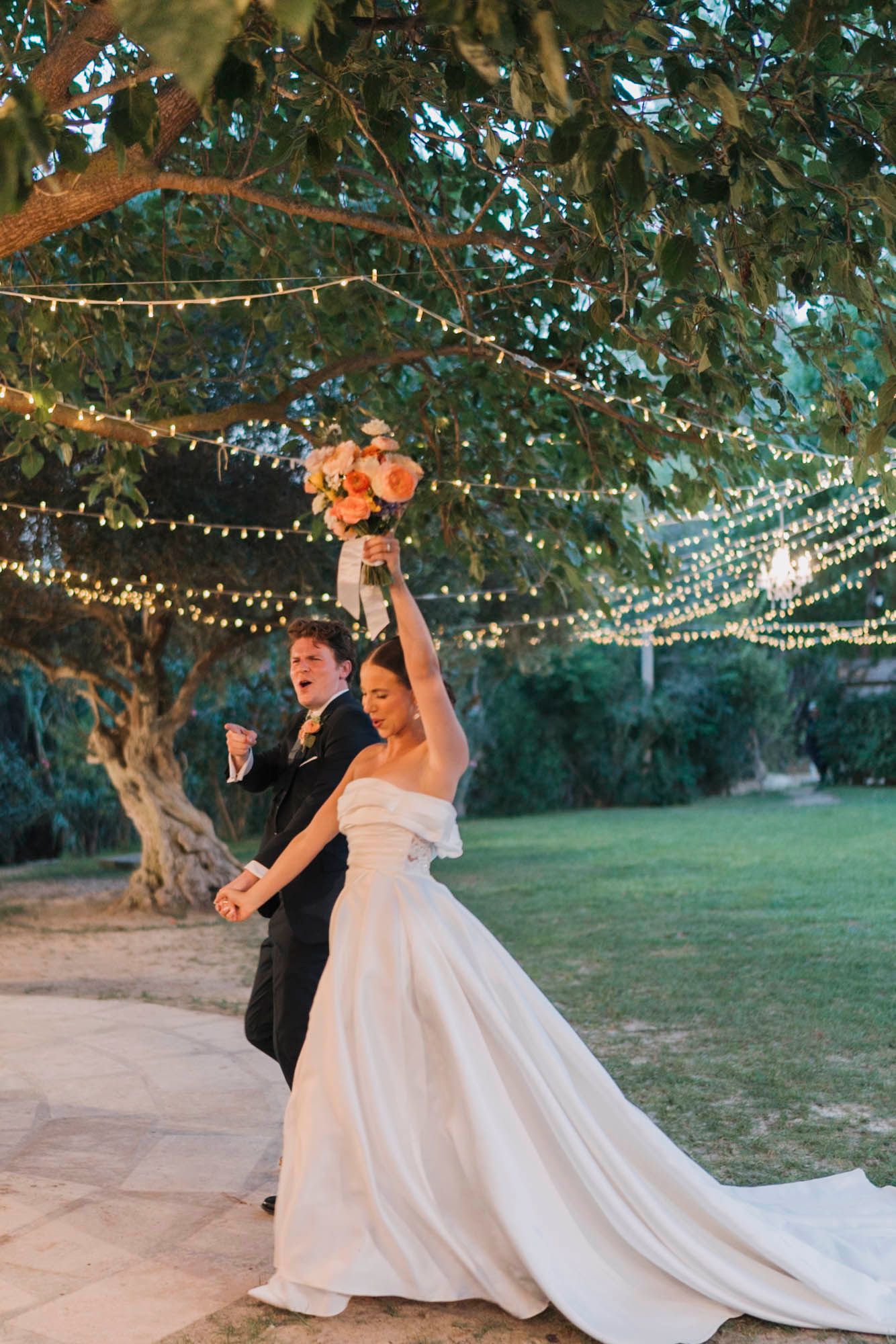 A couple celebrates during their outdoor wedding reception beneath canopies of string lights and tree branches.