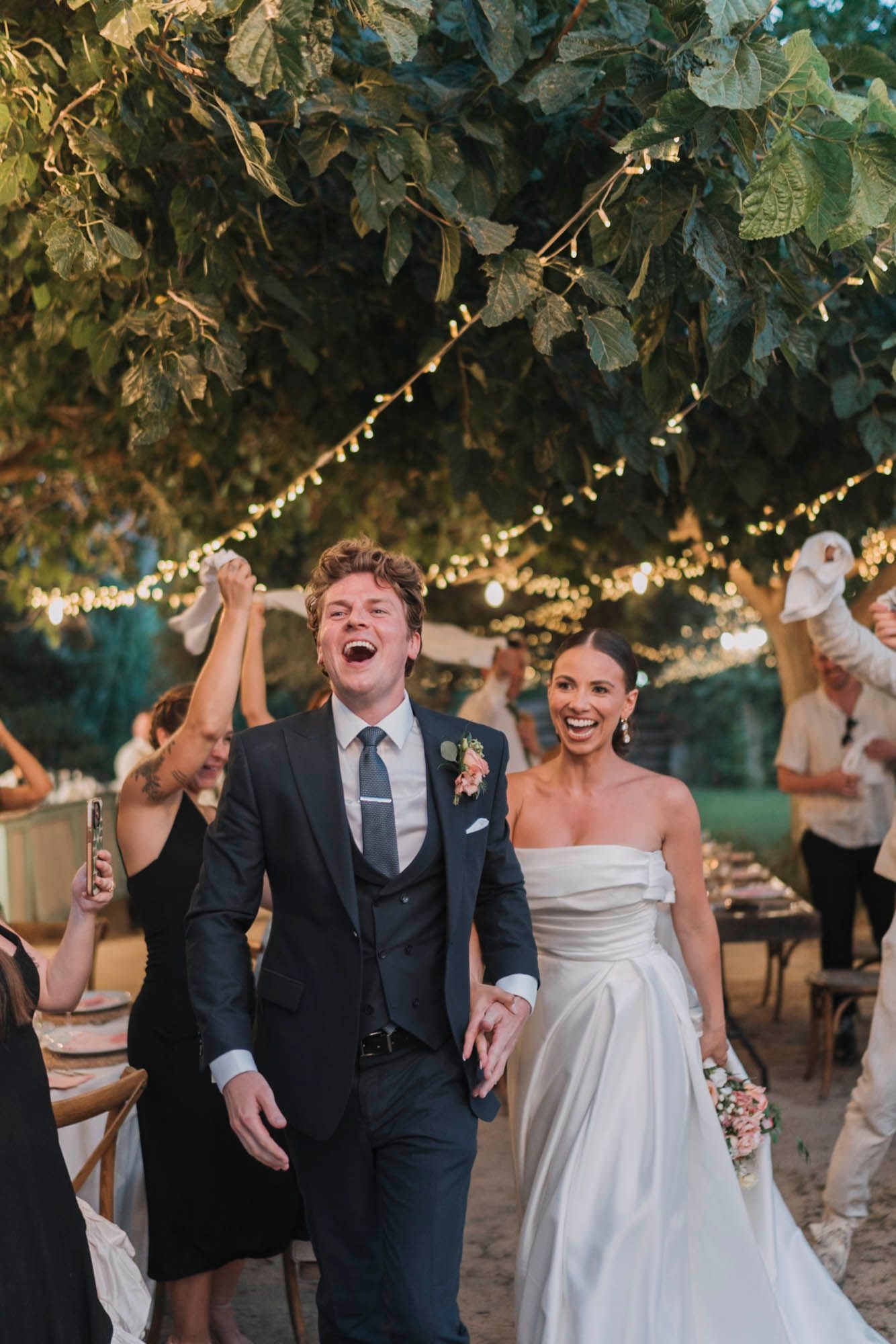 Joyful celebration at an outdoor wedding reception under string lights and trees.