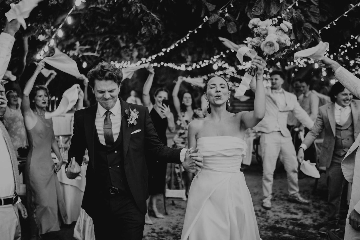 Black and white photo of wedding celebration with guests raising their arms under string lights.
