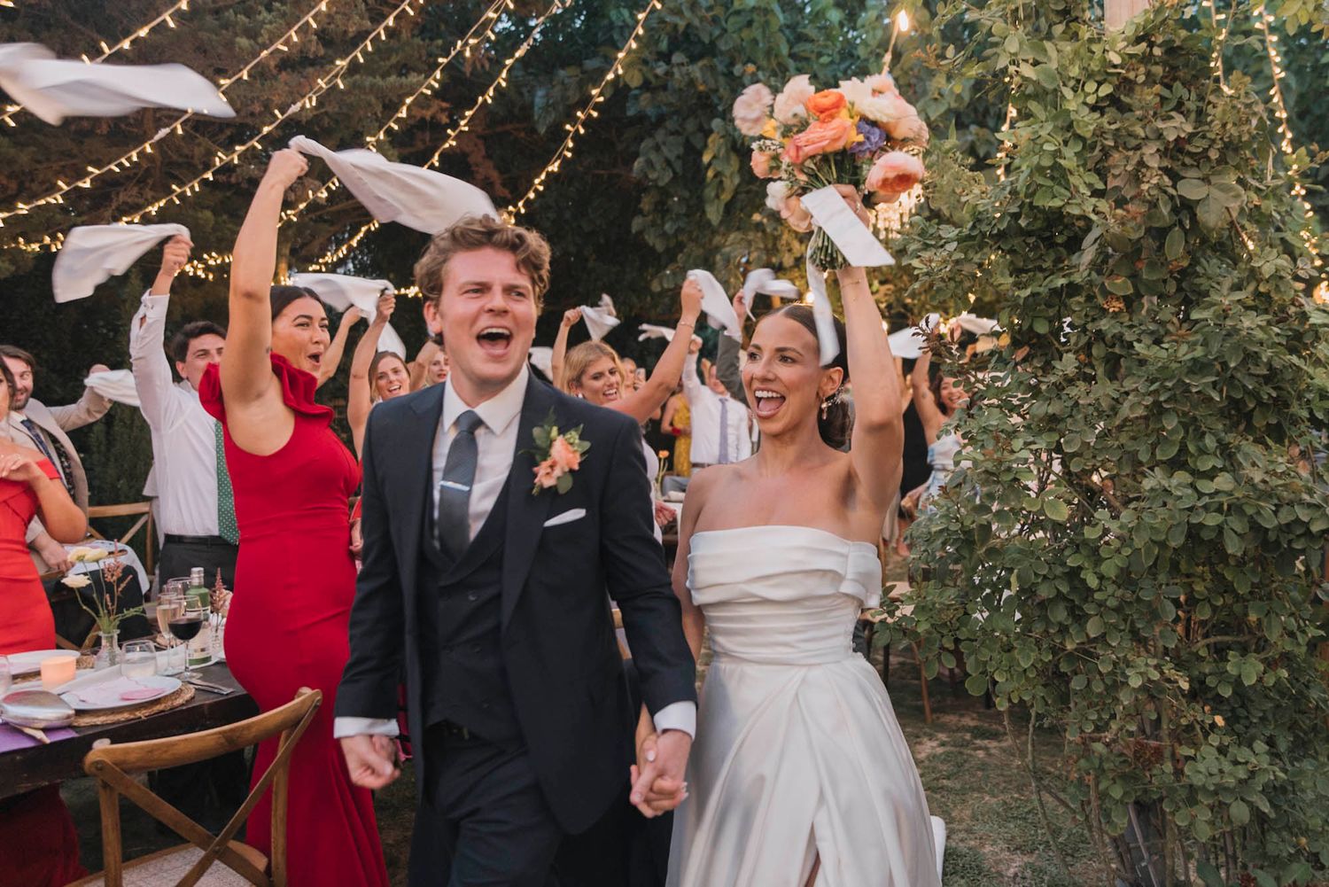 Wedding celebration under string lights with guests cheering and raising their arms in celebration.